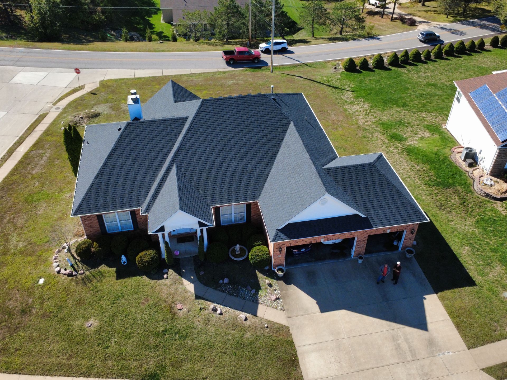 Aerial view of a residential house with a dark, textured roof, brick siding, and a driveway in a suburban neighborhood.