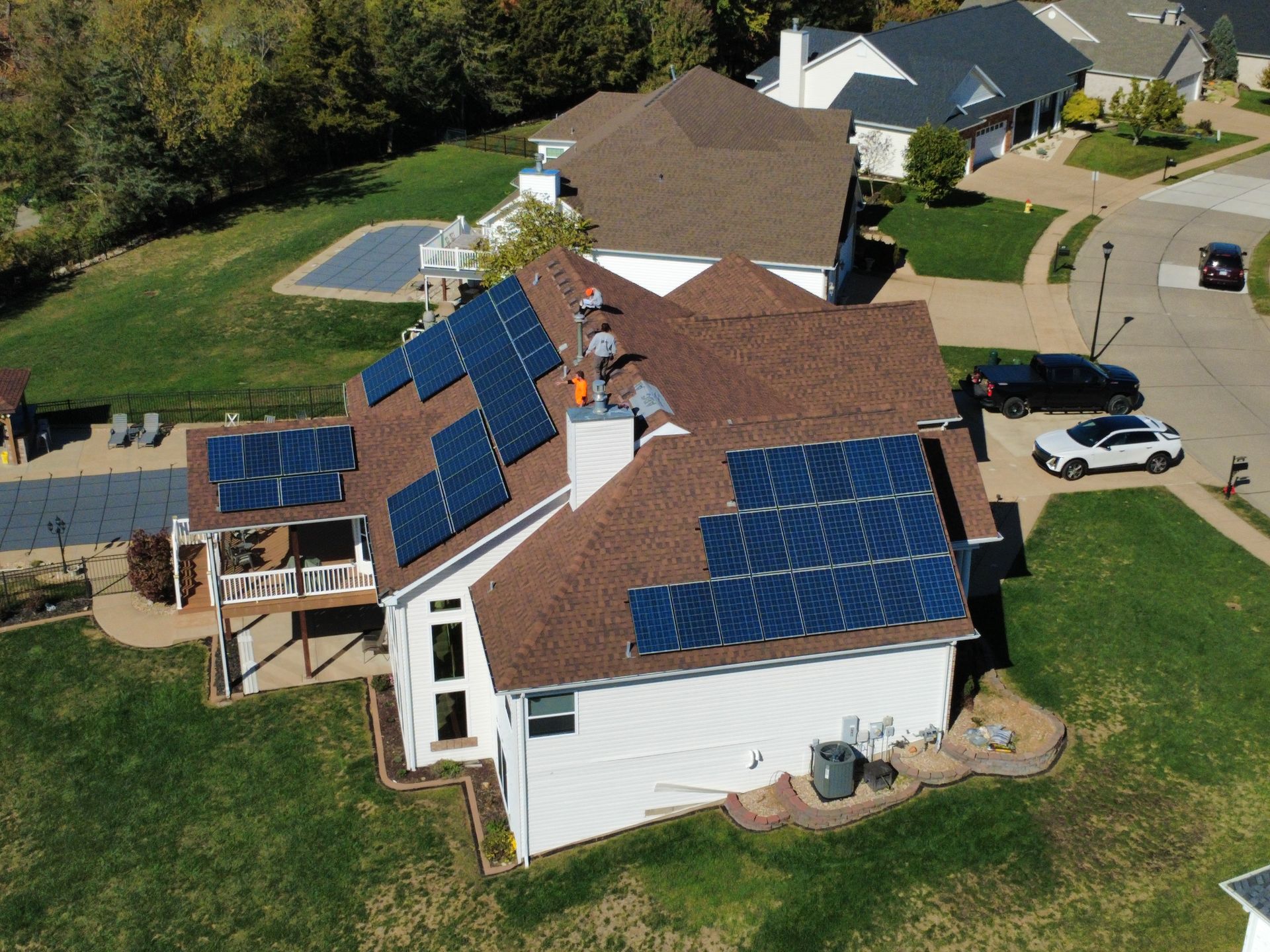 Aerial view of a residential house with a brown shingled roof, featuring solar panel arrays and two workers on the roof.