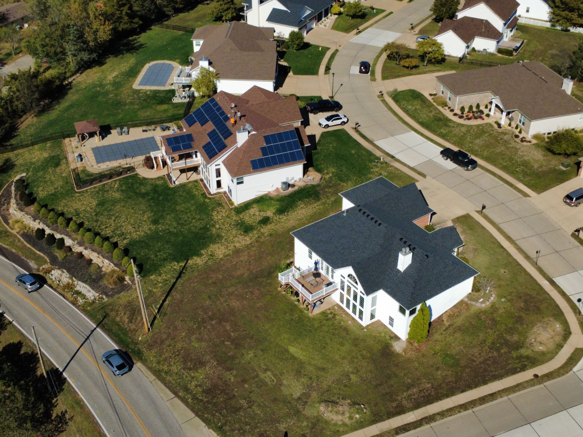 Aerial view of suburban houses with lawns and swimming pools, set along winding roads on a sunny day.