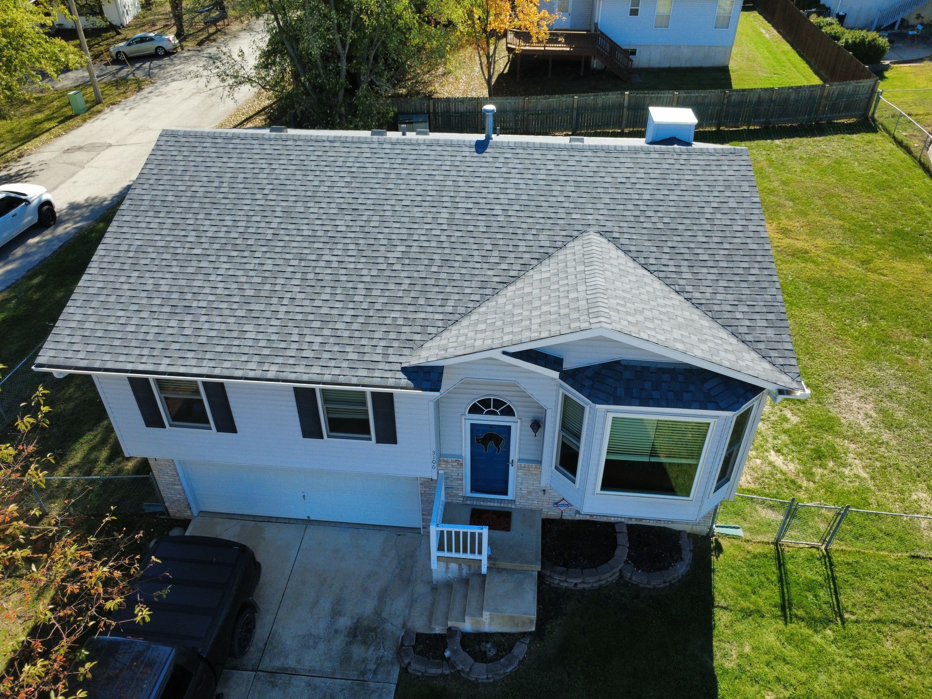 An elevated, aerial view of a single-story, white suburban house with a gray shingled roof, a blue front door, and a yard.