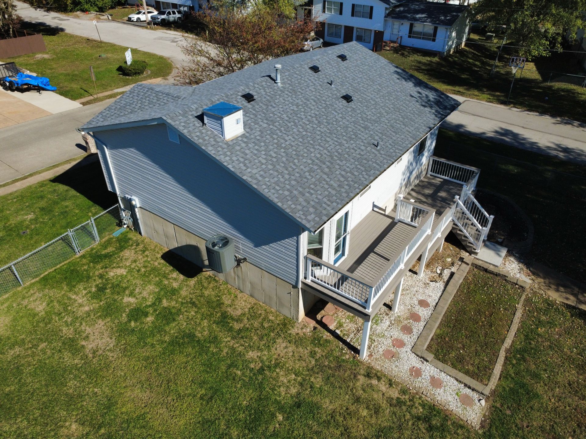 Aerial view of a gray-roofed residential house with a back deck, a patio area, and a green lawn in a suburban setting.
