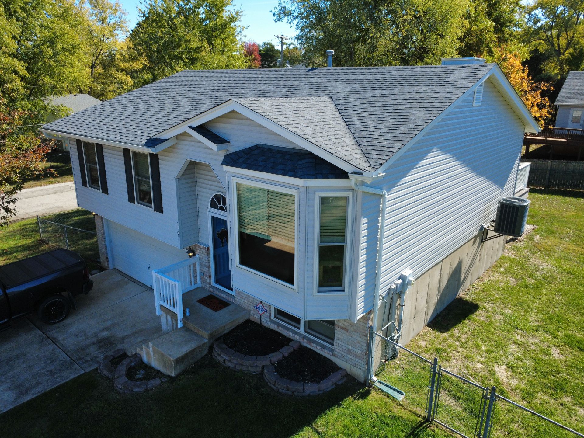 Aerial view of a light-colored, split-level suburban house with a gray shingled roof, front bay window, and attached garage.