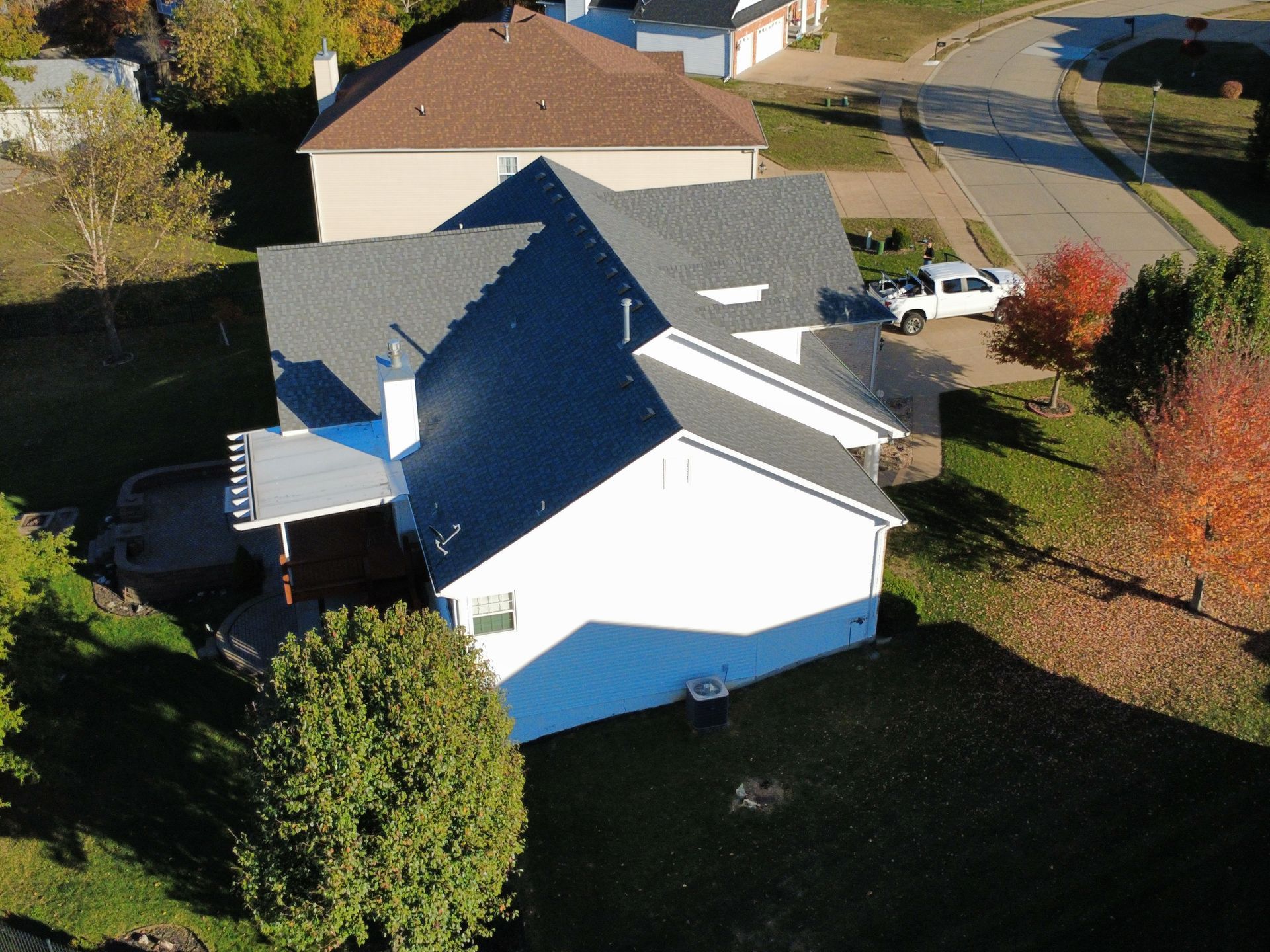 Aerial view of a residential suburban house with a gray shingled roof, white siding, and a backyard patio.