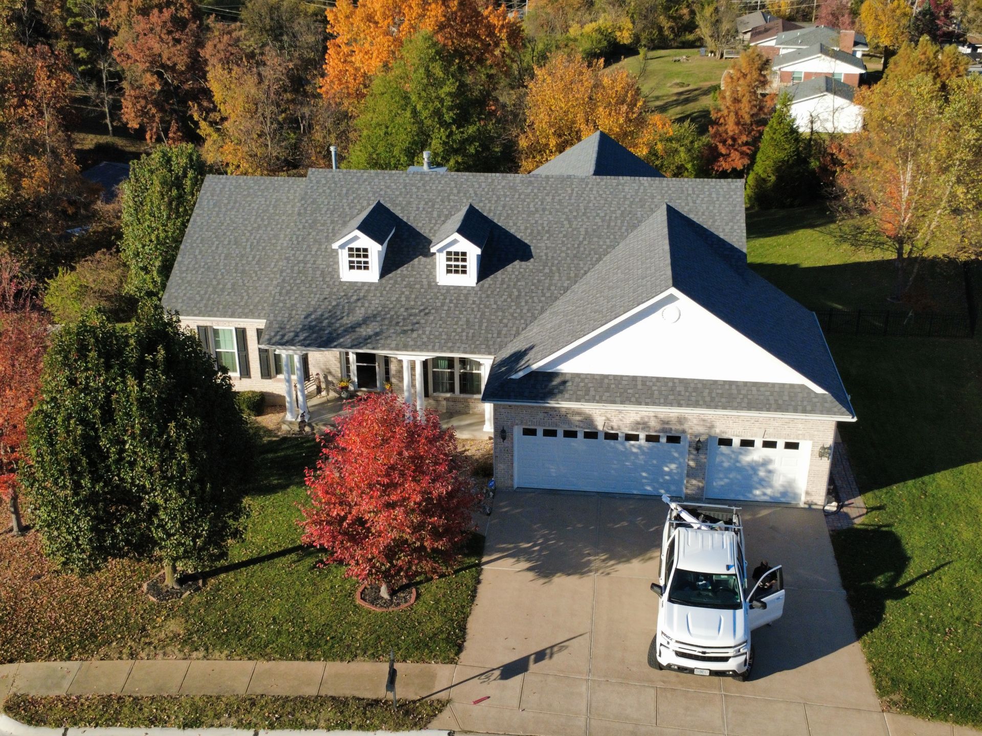 Aerial view of a house with a gray roof and a white garage, featuring a service truck parked in the driveway in autumn.