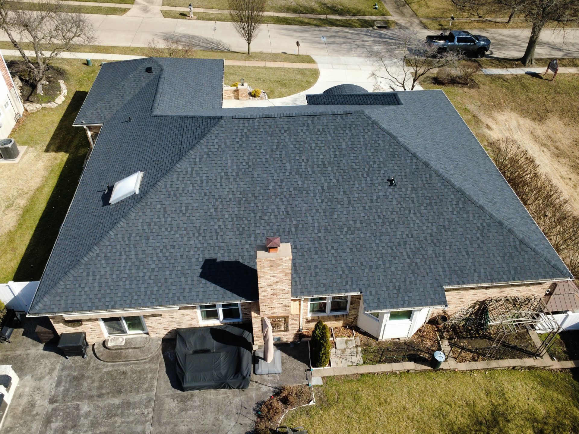 An aerial view of a brick house with a dark shingled roof, featuring a brick chimney and a backyard patio area.