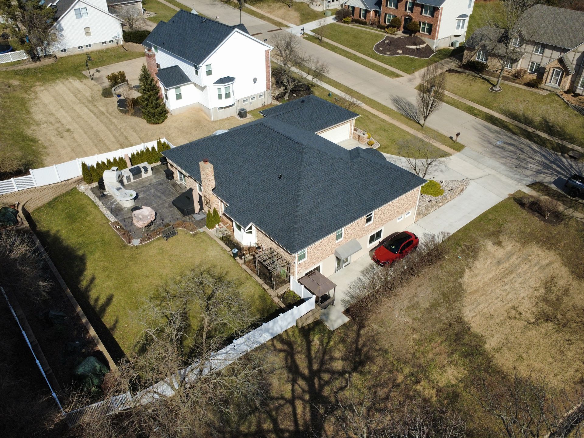 Aerial view of a residential house with a dark shingled roof, a stone patio, and a red car parked on the concrete driveway.