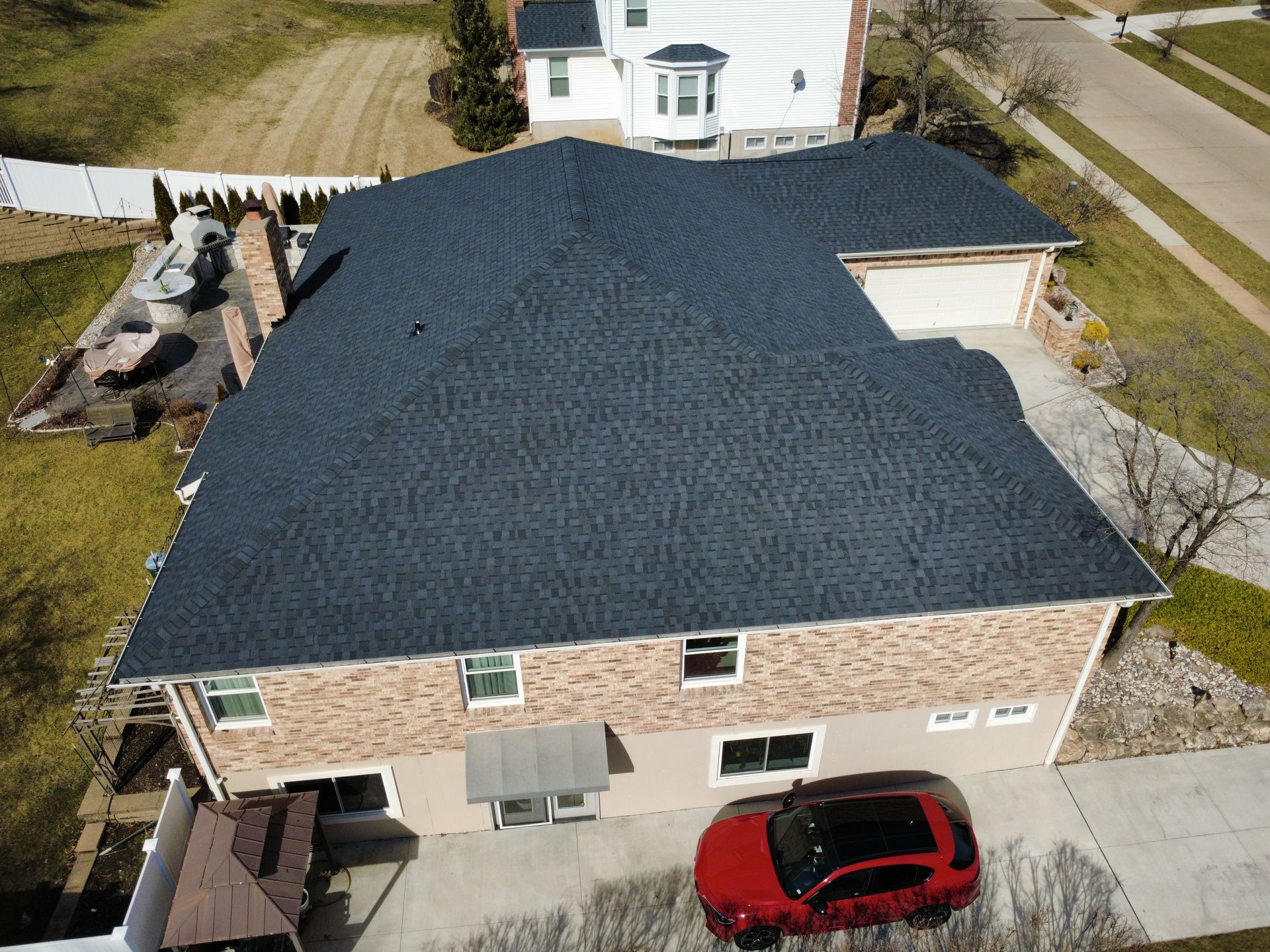 An aerial view of a brick house with a dark shingled roof, featuring a red car parked on the concrete driveway.