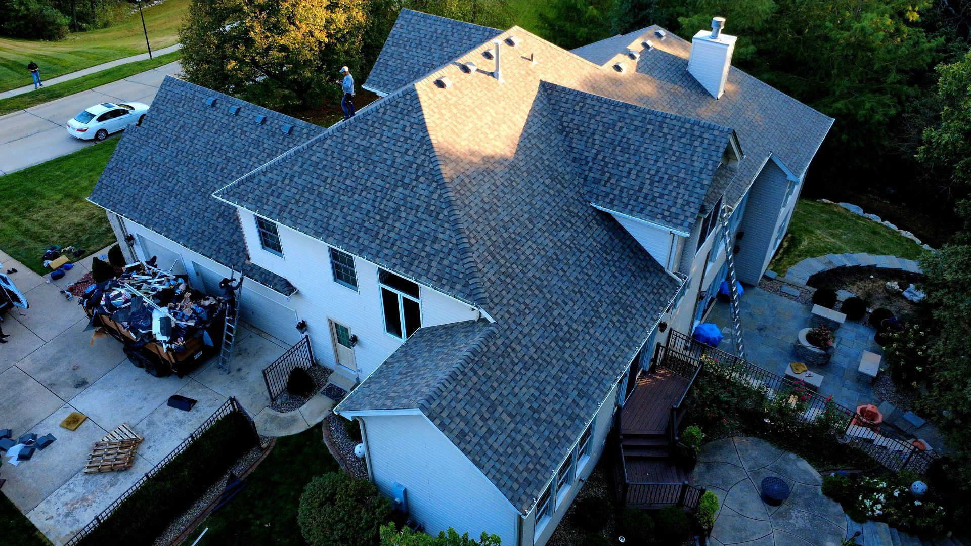 Aerial view of a residential house with a grey shingled roof, a chimney, and a paved patio area in a suburban setting.