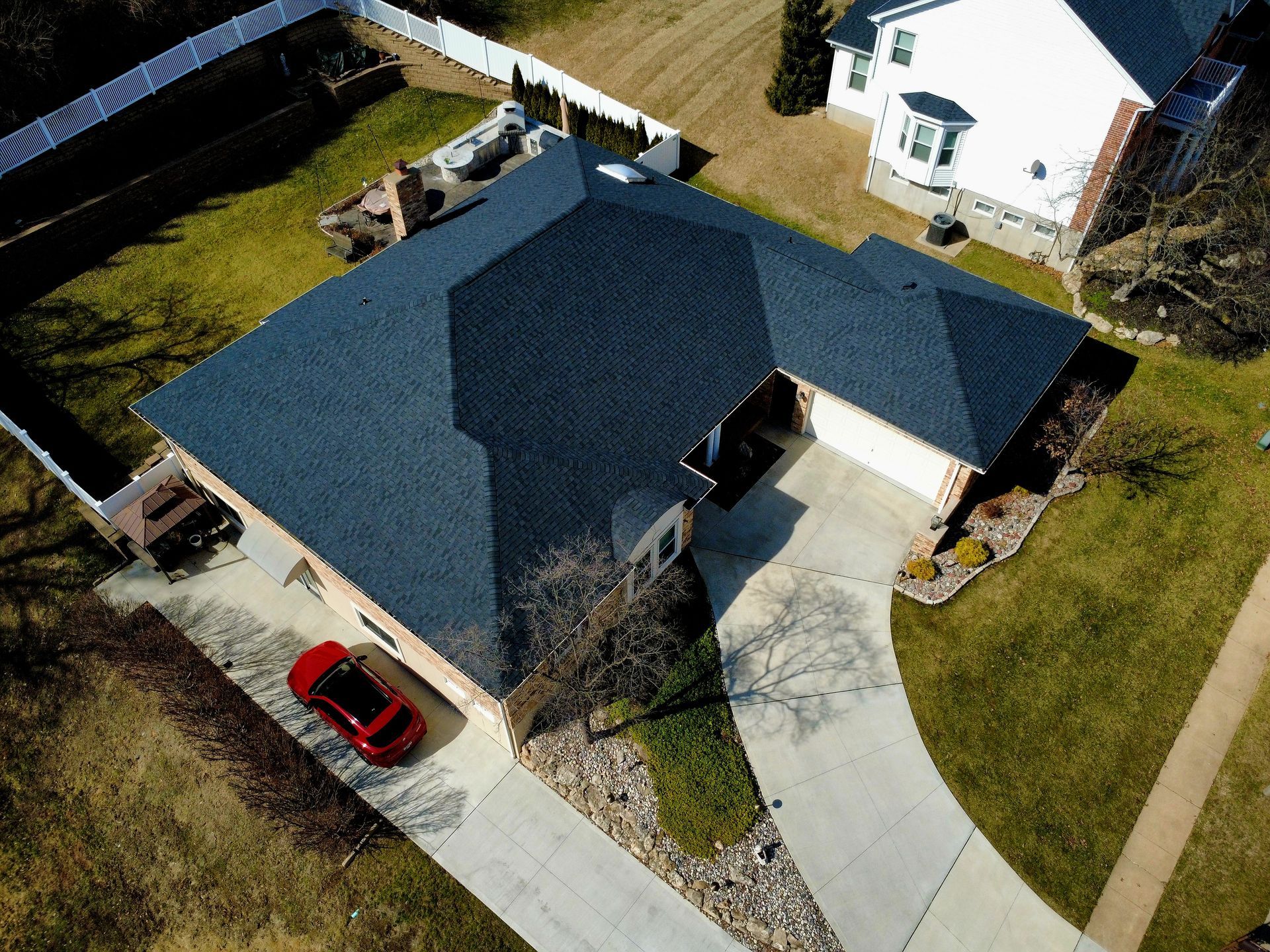 Aerial view of a residential house with a dark shingled roof, concrete driveway, and a red car parked on the side.