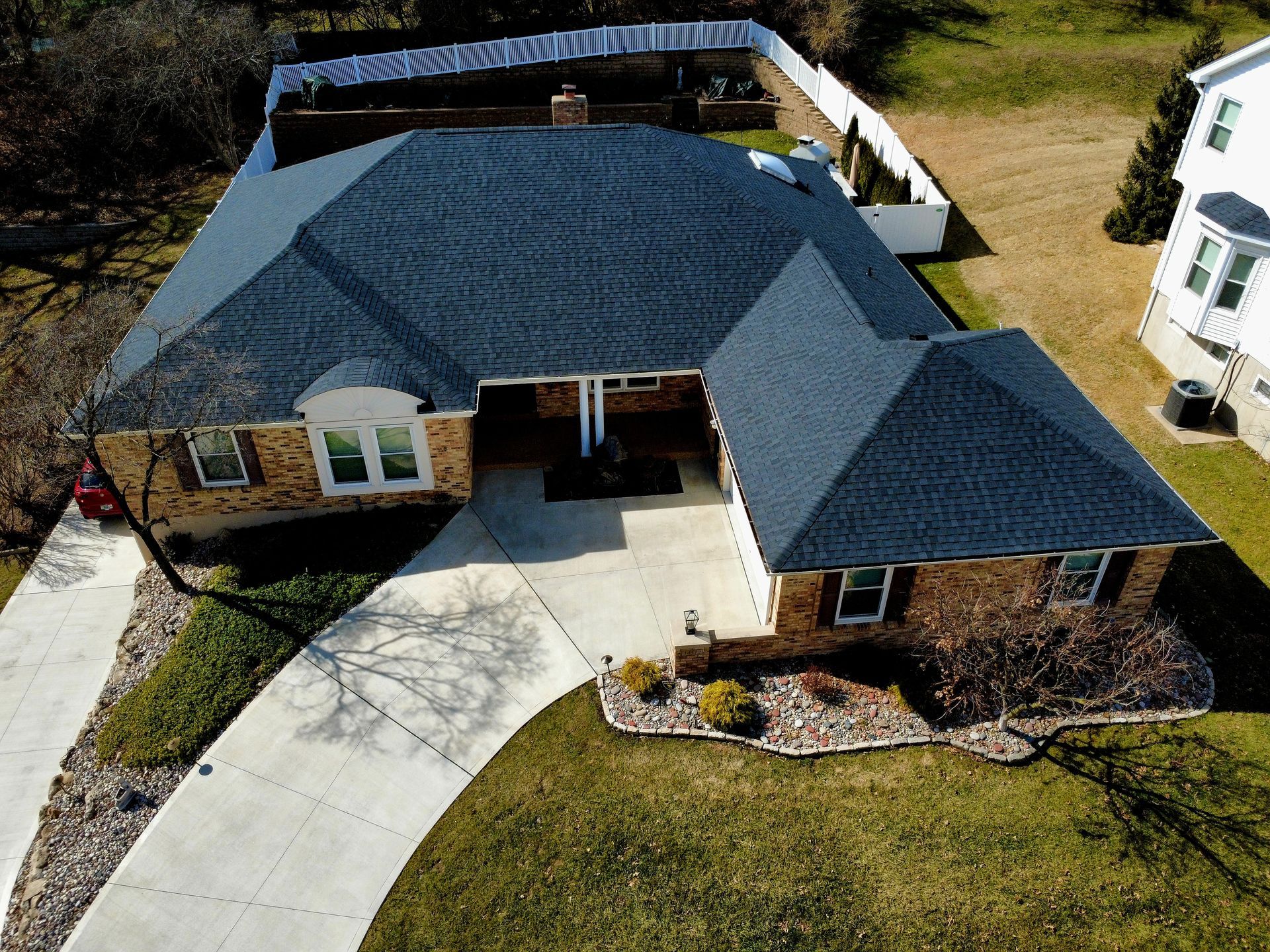 Aerial view of a suburban brick house with a dark shingled roof, a paved driveway, and a fenced backyard.