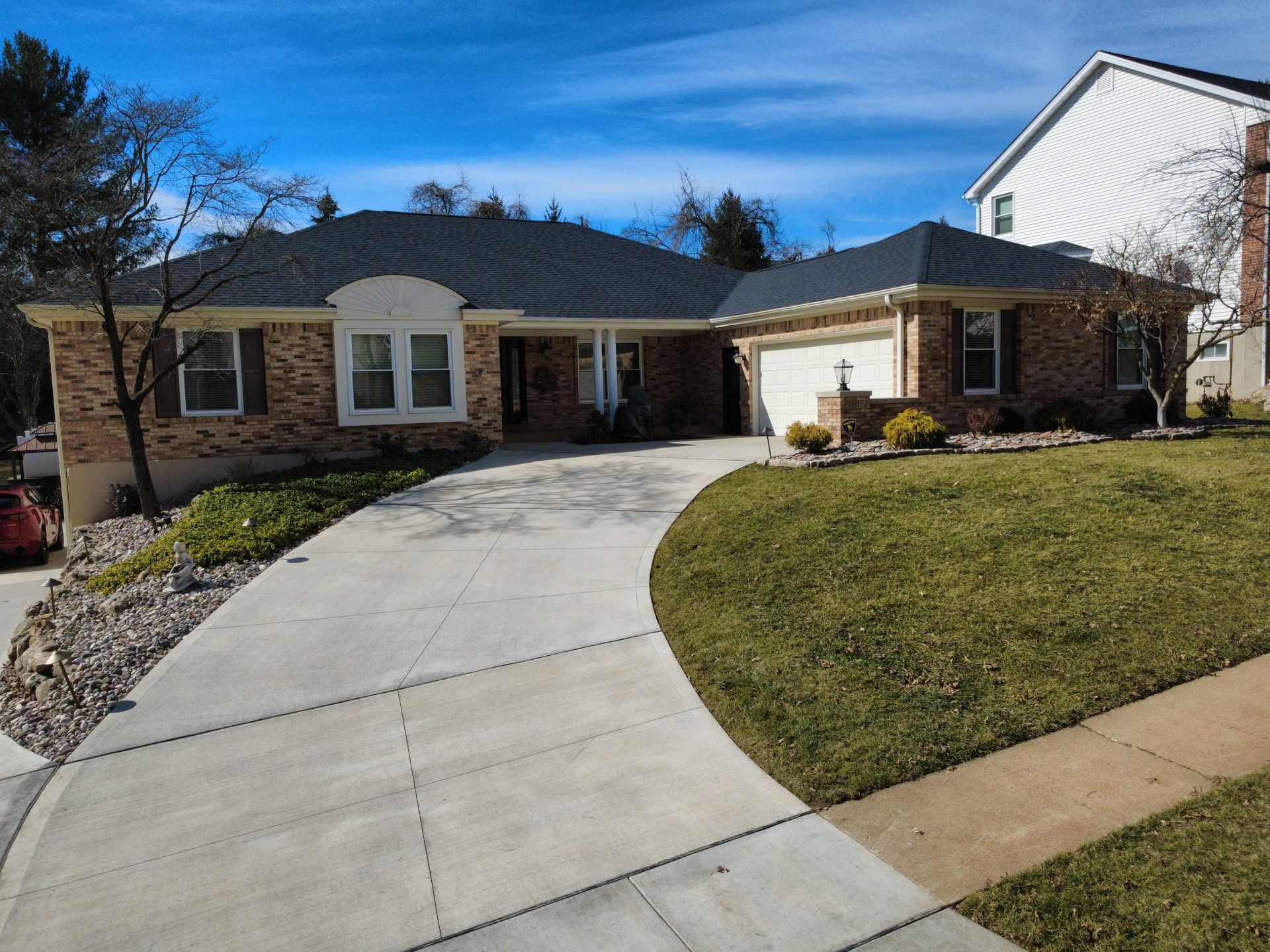 A one-story brick house with a dark roof and a concrete driveway, set in a suburban neighborhood on a sunny day.
