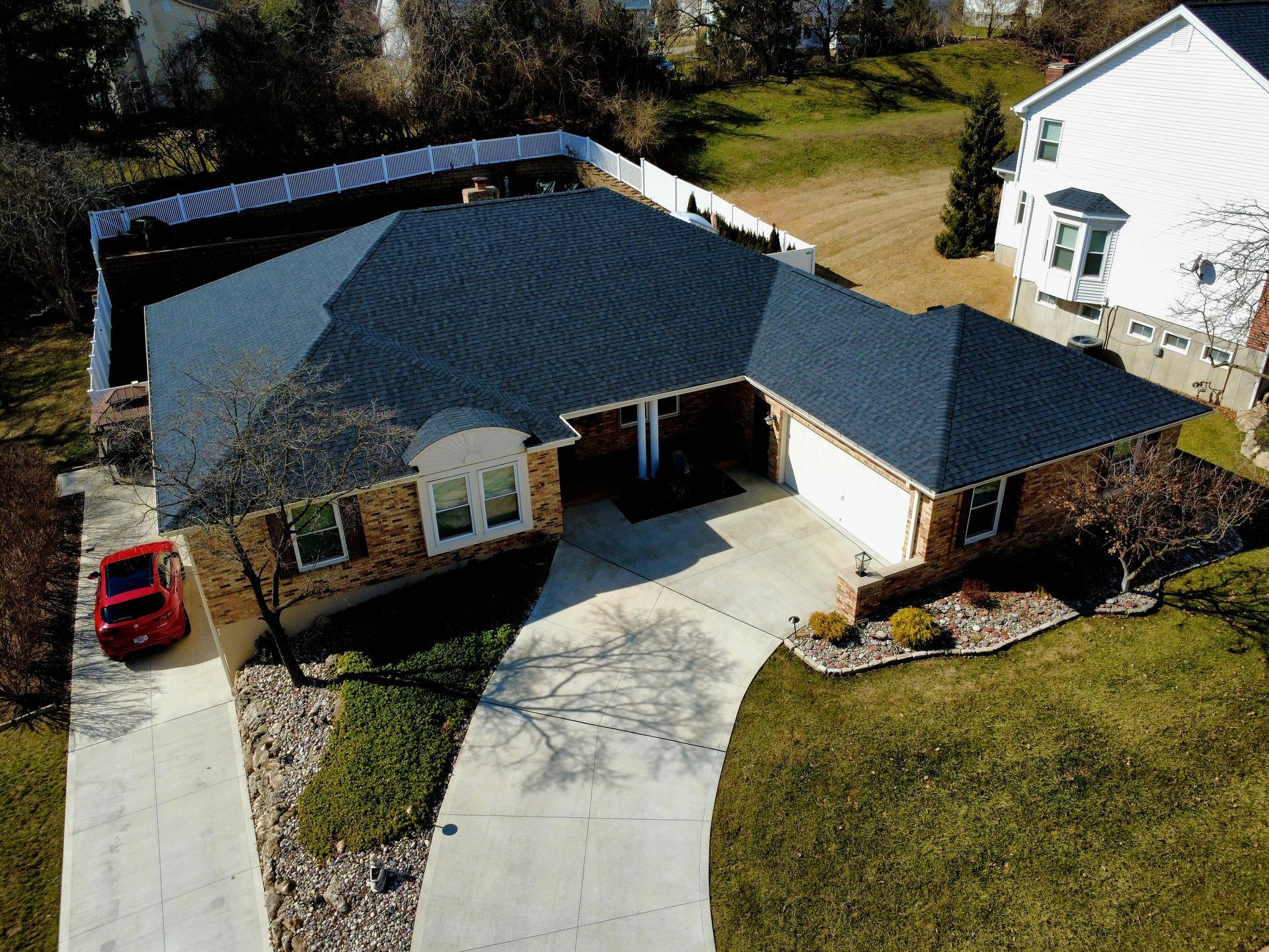An aerial view of a brick house with a dark roof and white trim, featuring a concrete driveway and a red car parked.