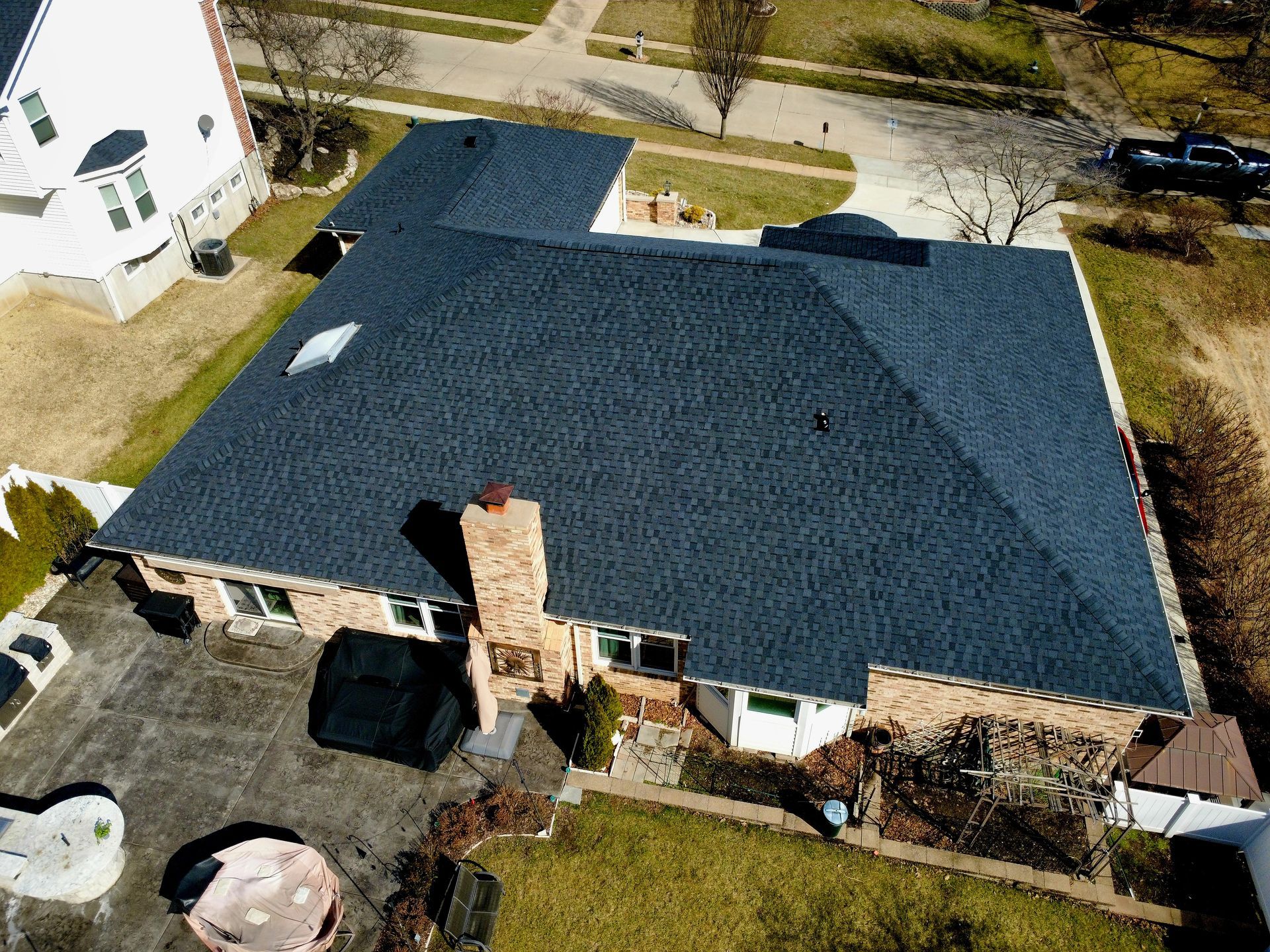 Aerial view of a residential brick house with a newly installed dark shingle roof, paved patio, and suburban backyard.