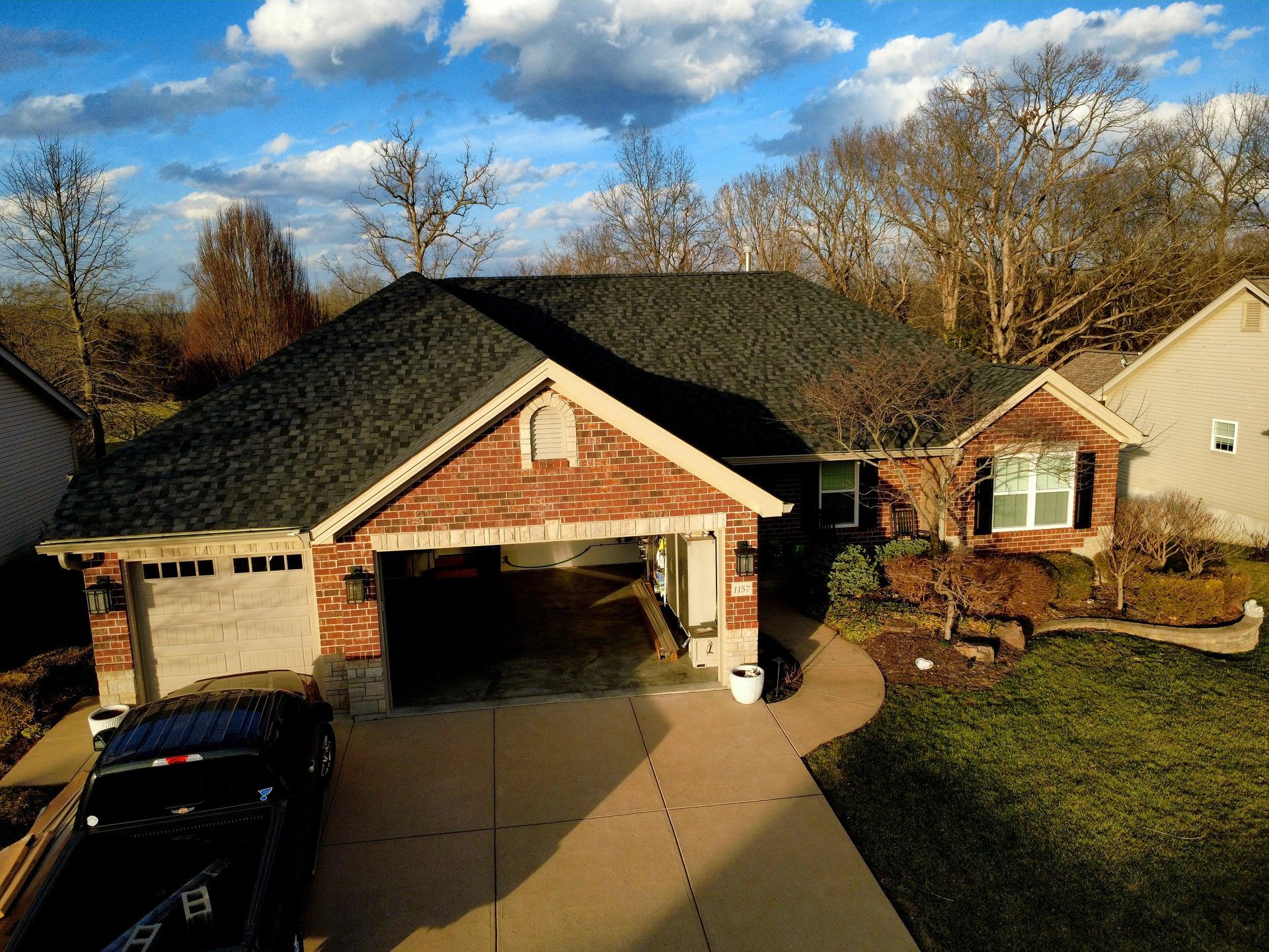 An aerial view of a single-story brick house with a dark shingled roof, a two-car garage, and a driveway in a suburban area.