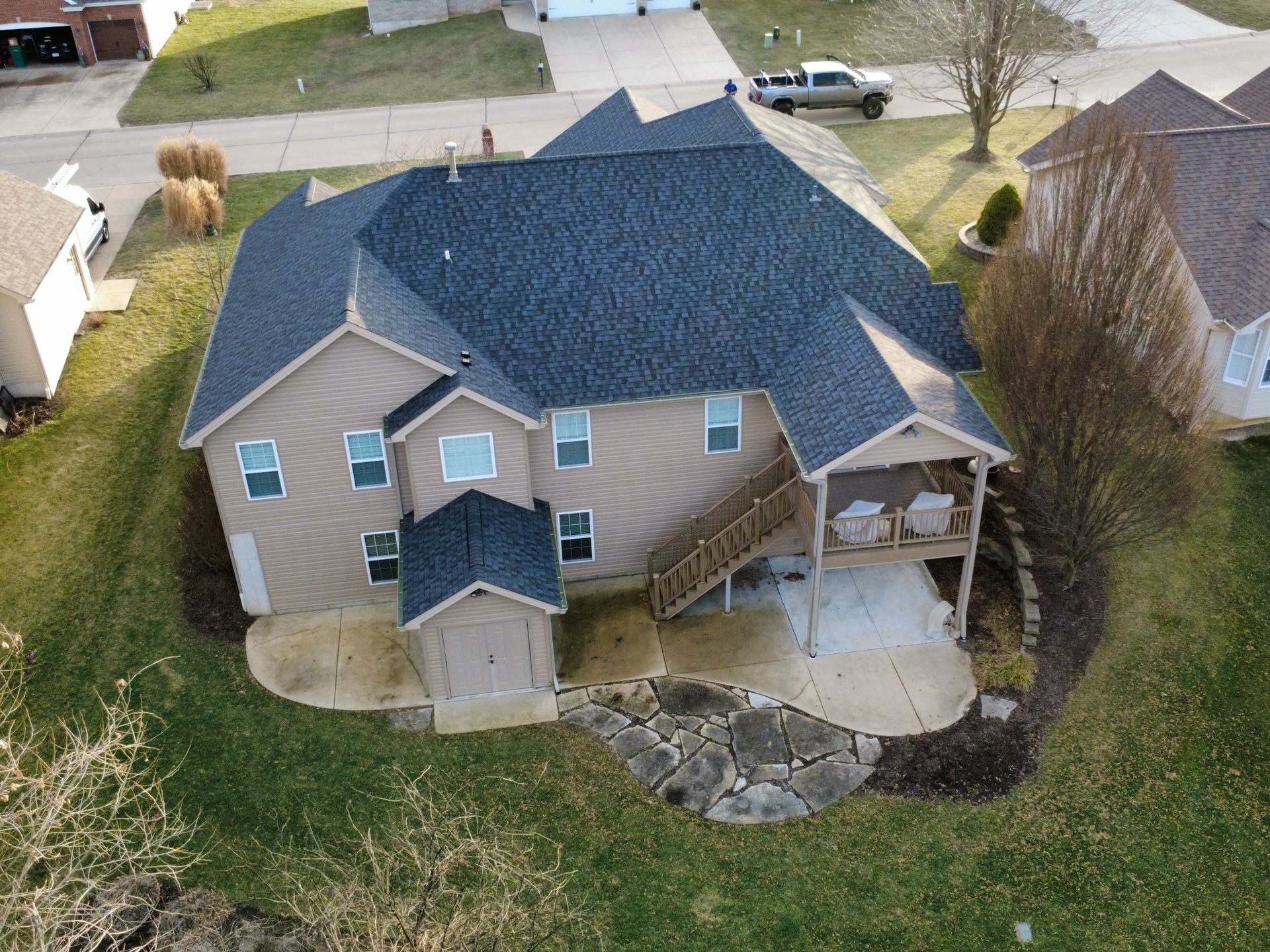 Aerial view of a two-story tan suburban house with a dark gray shingled roof, a back deck, and a stone patio.