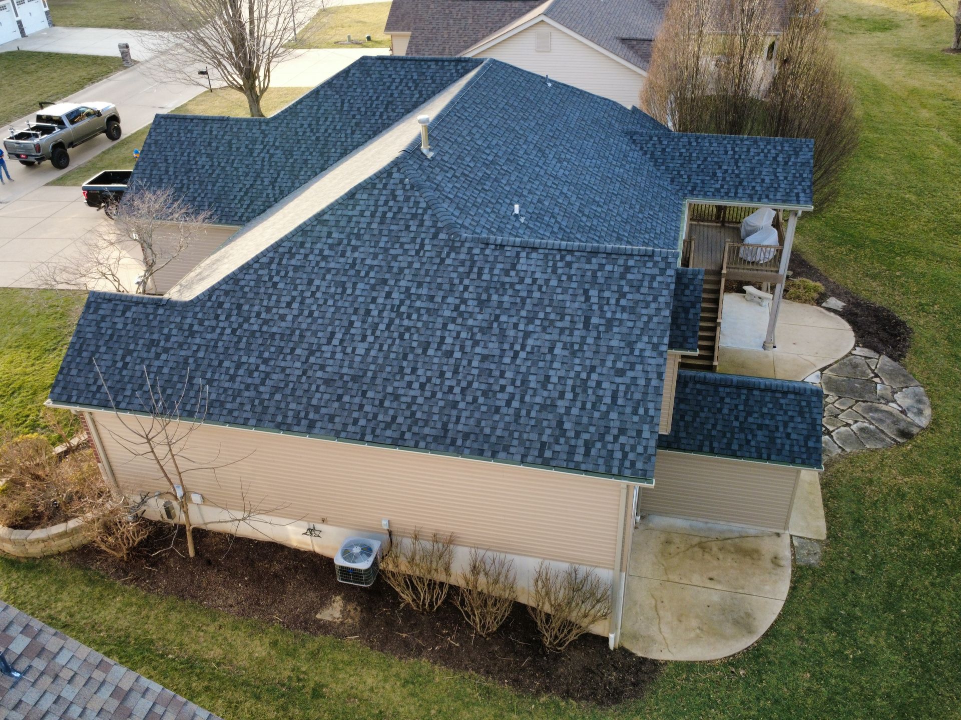 An aerial view of a tan house with a dark blue shingled roof, a stone patio, and surrounding lawn.