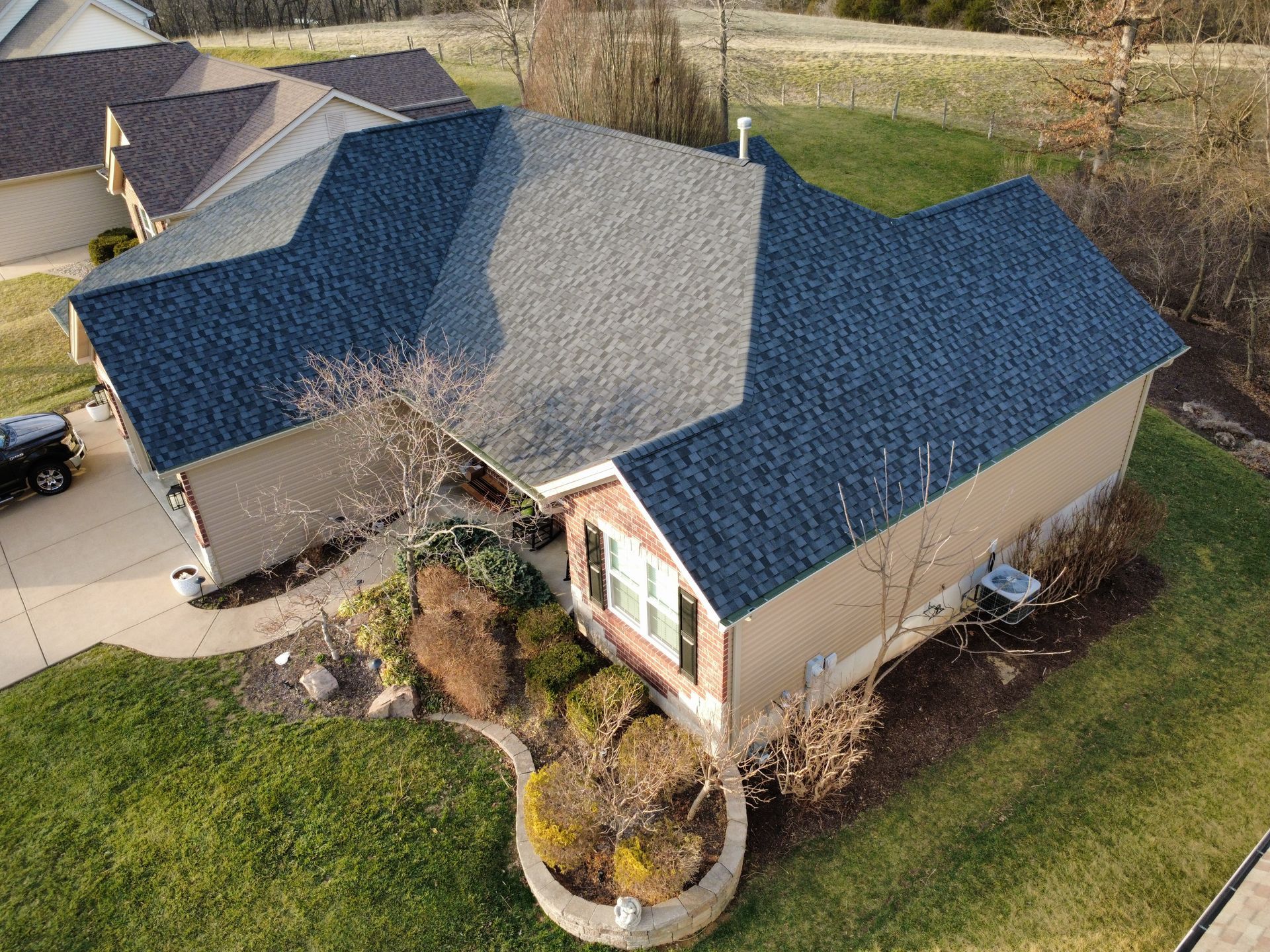 High-angle aerial view of a suburban house with a dark blue shingled roof, tan siding, and a curved stone landscape bed.