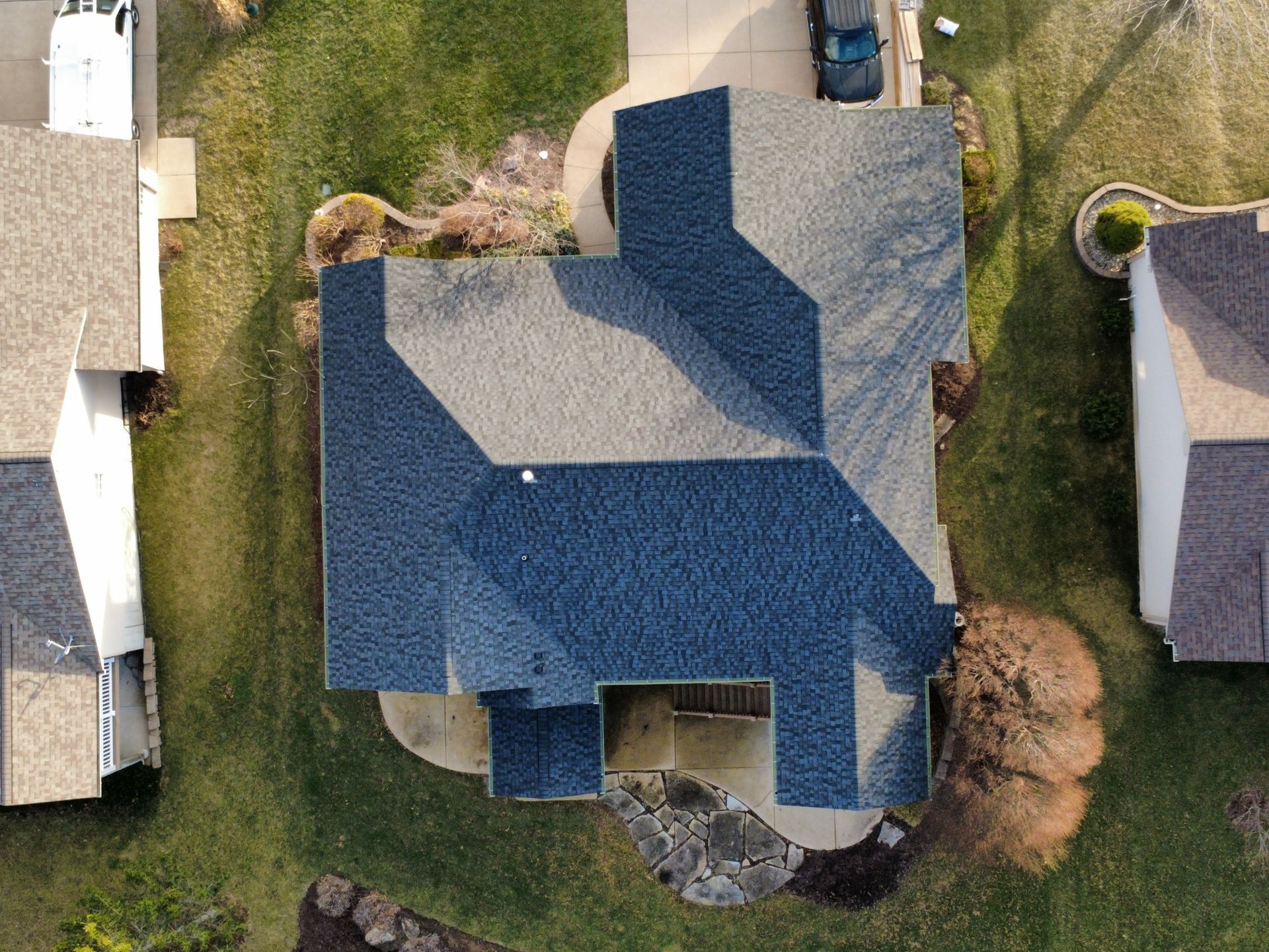 Aerial view of a residential house with a blue and grey shingled roof surrounded by a lawn and adjacent properties.