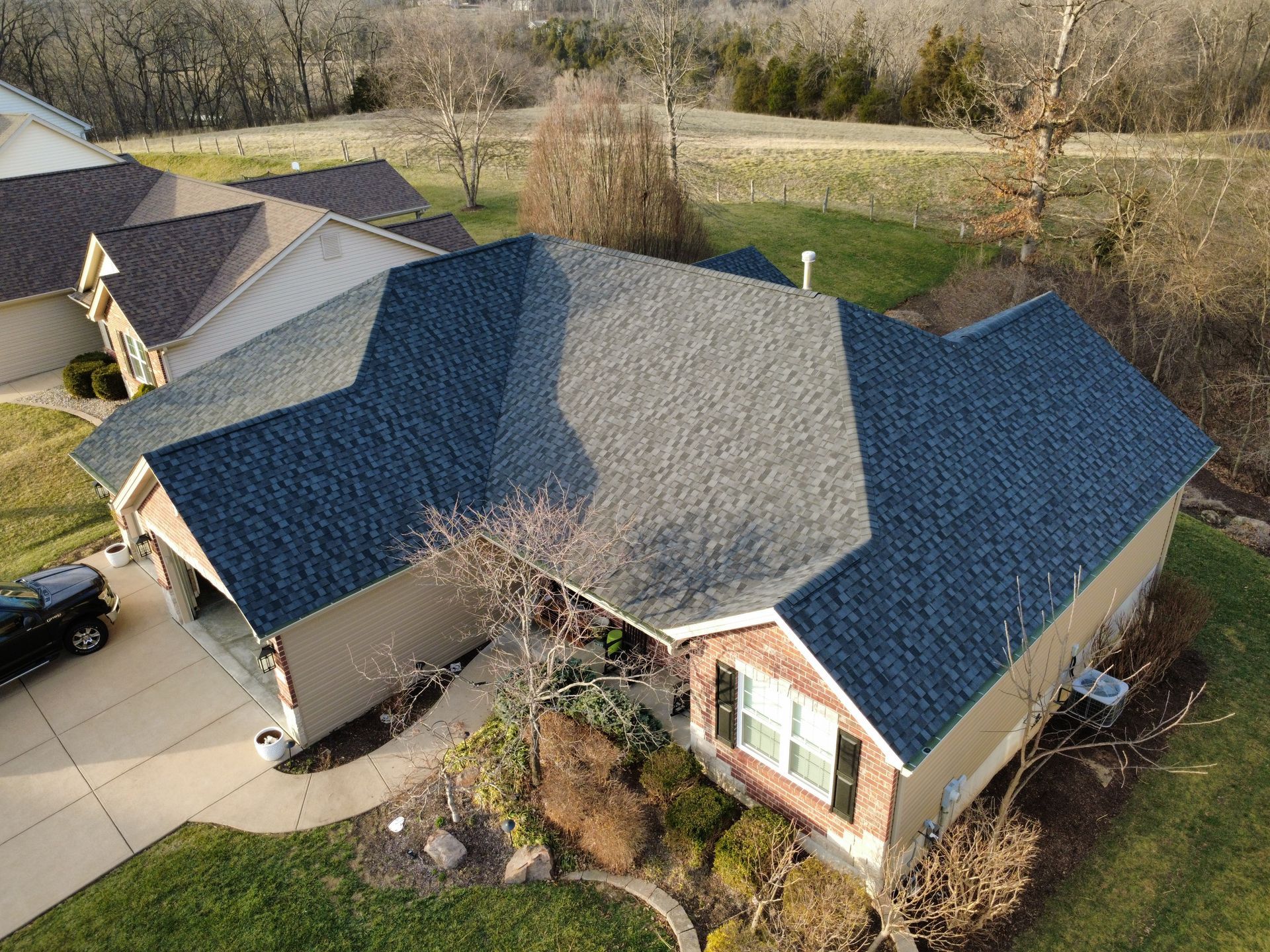 Aerial view of a residential suburban house with dark gray asphalt shingled roofing and a paved driveway.