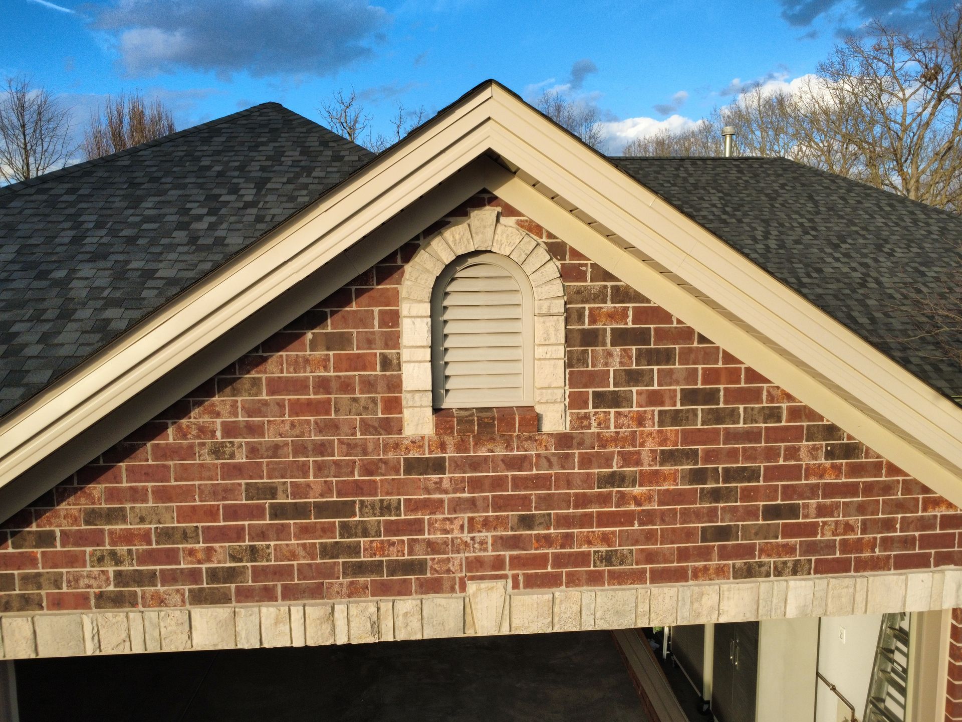 A red brick gable end with a light-colored arched vent and stone trim above a garage roof against a blue sky.