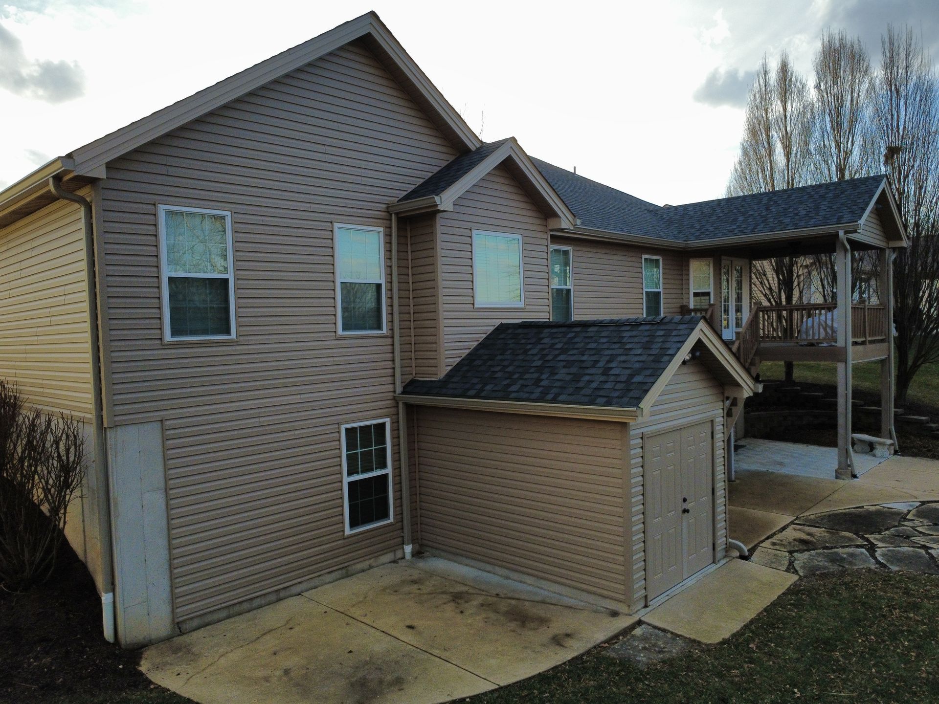 A tan two-story house with a small shed addition, a raised wooden deck, and a concrete patio in the backyard.