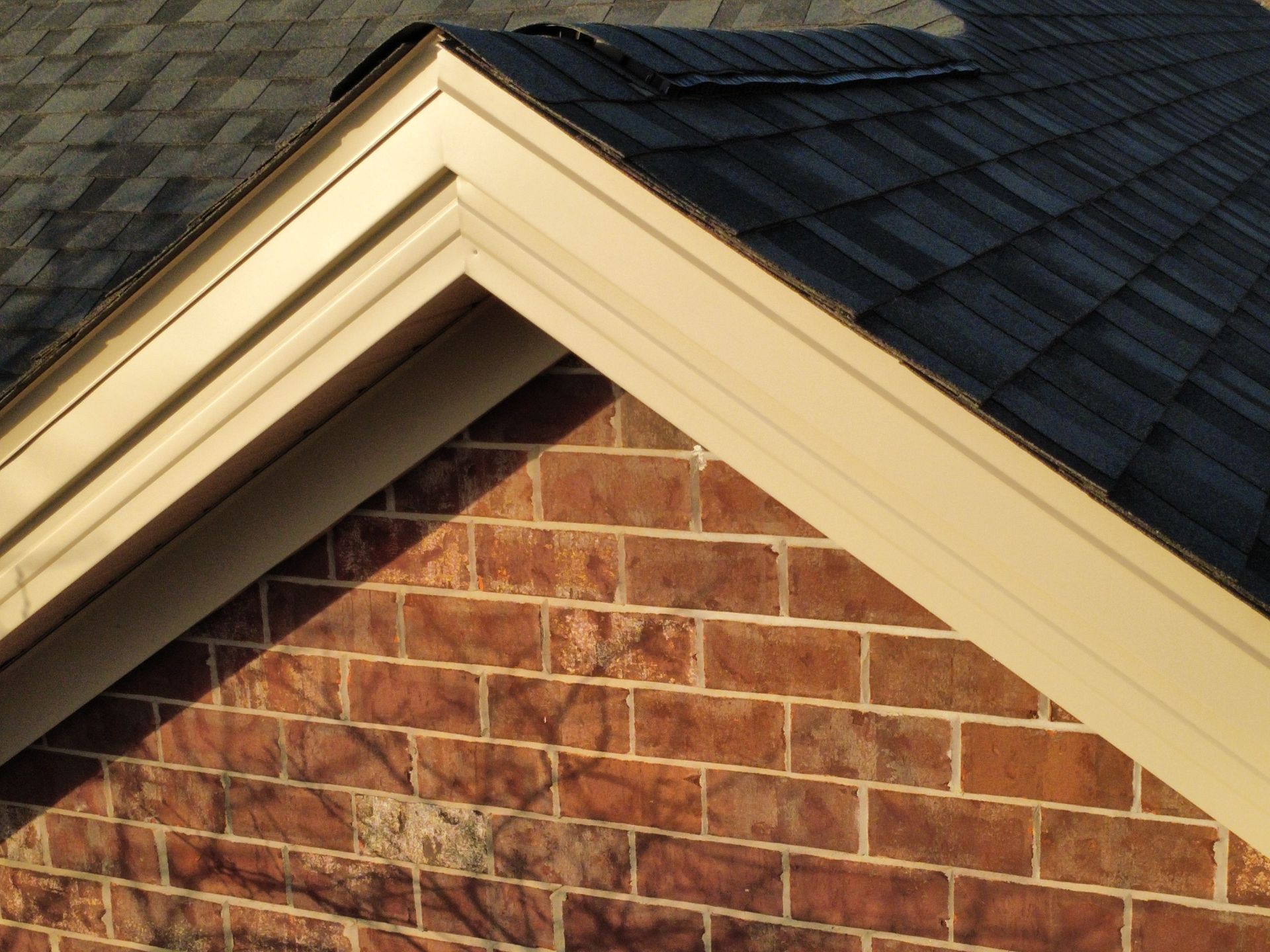 Close-up of a house gable featuring tan trim, dark grey asphalt shingles, and red brick siding.