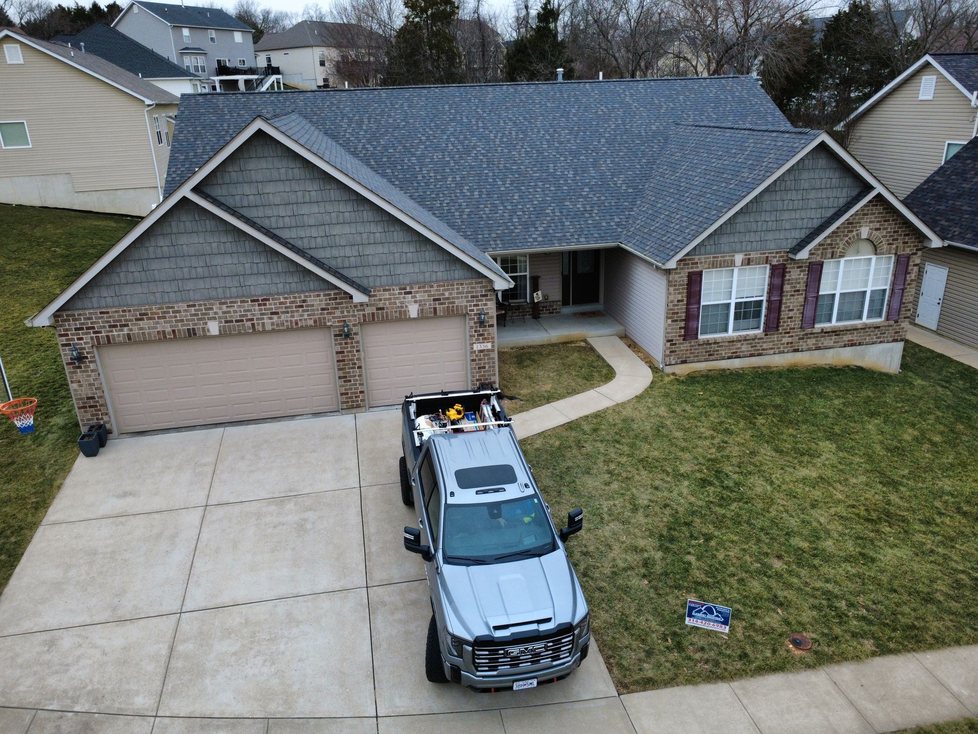 An aerial view of a single-story brick house with a gray roof and a silver pickup truck parked in the driveway.