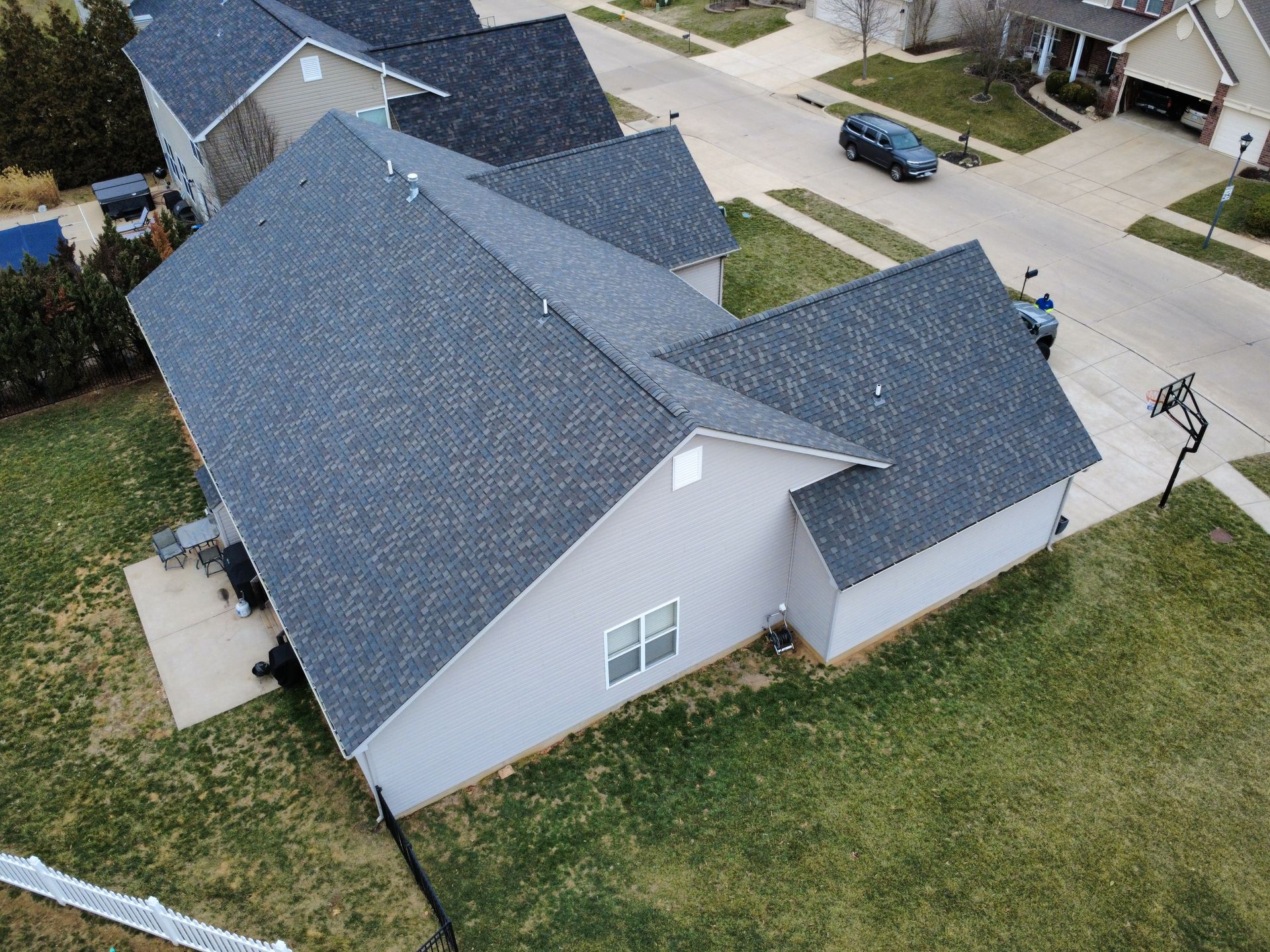 An aerial view of a gray residential house with a dark shingled roof, a concrete driveway, and surrounding green lawn.
