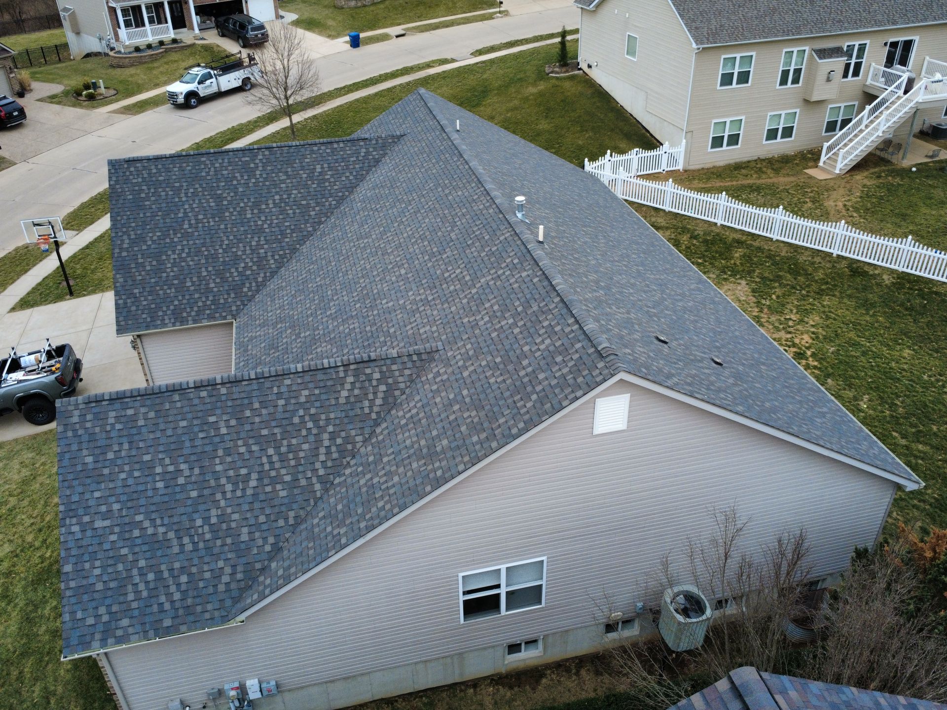 An aerial view of a tan-sided suburban house with a dark shingled roof, next to a paved street and a neighboring home.