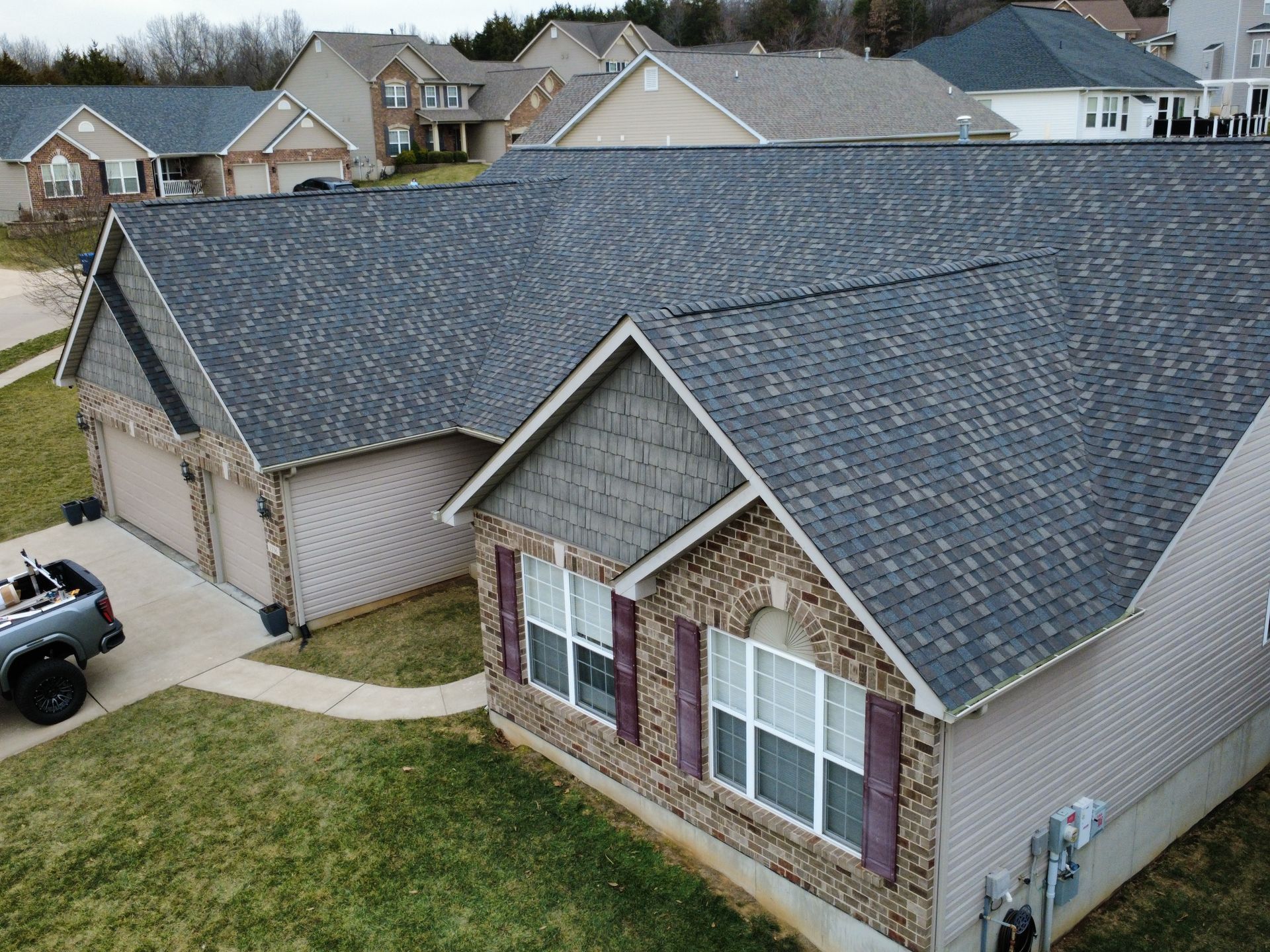 An aerial view of a residential house with a dark shingled roof, brick siding, and a two-car garage on a suburban street.