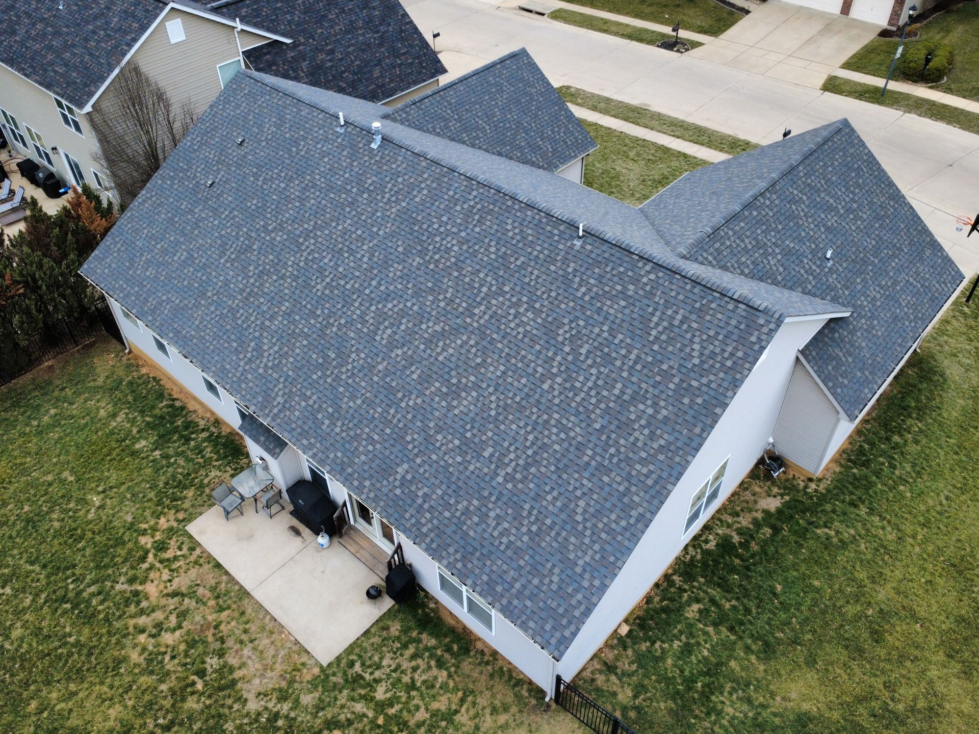An aerial view of a single-family home with a gray shingled roof, a small concrete patio, and a grassy backyard.