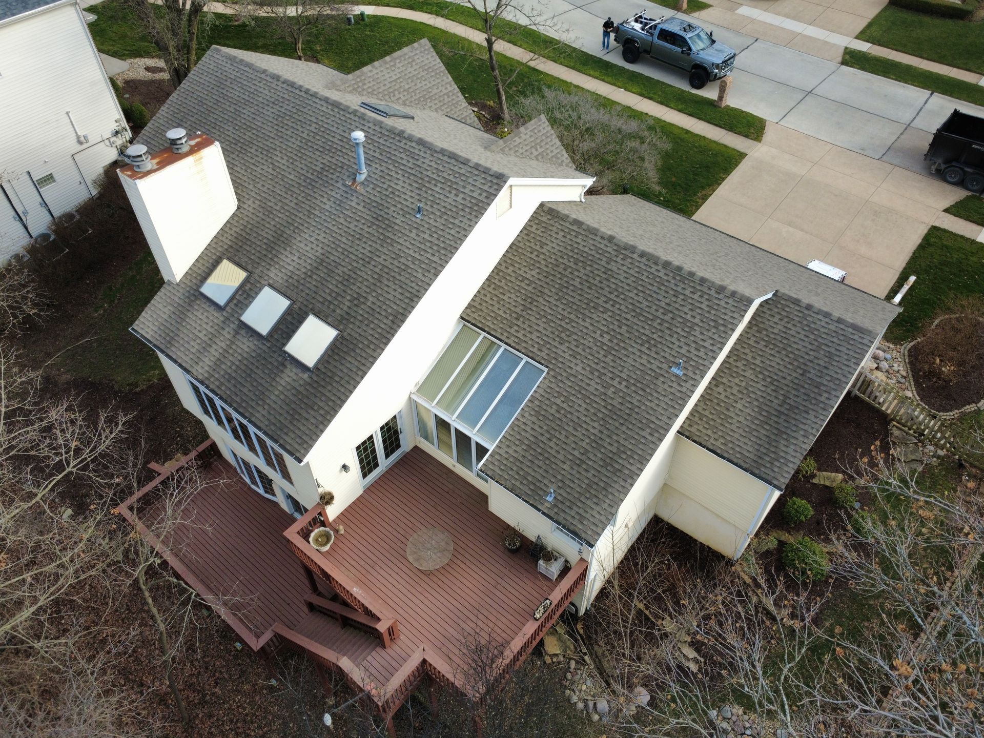 Aerial view of a two-story house with a wooden deck, gray roof shingles, three skylights, and a small sunroom.