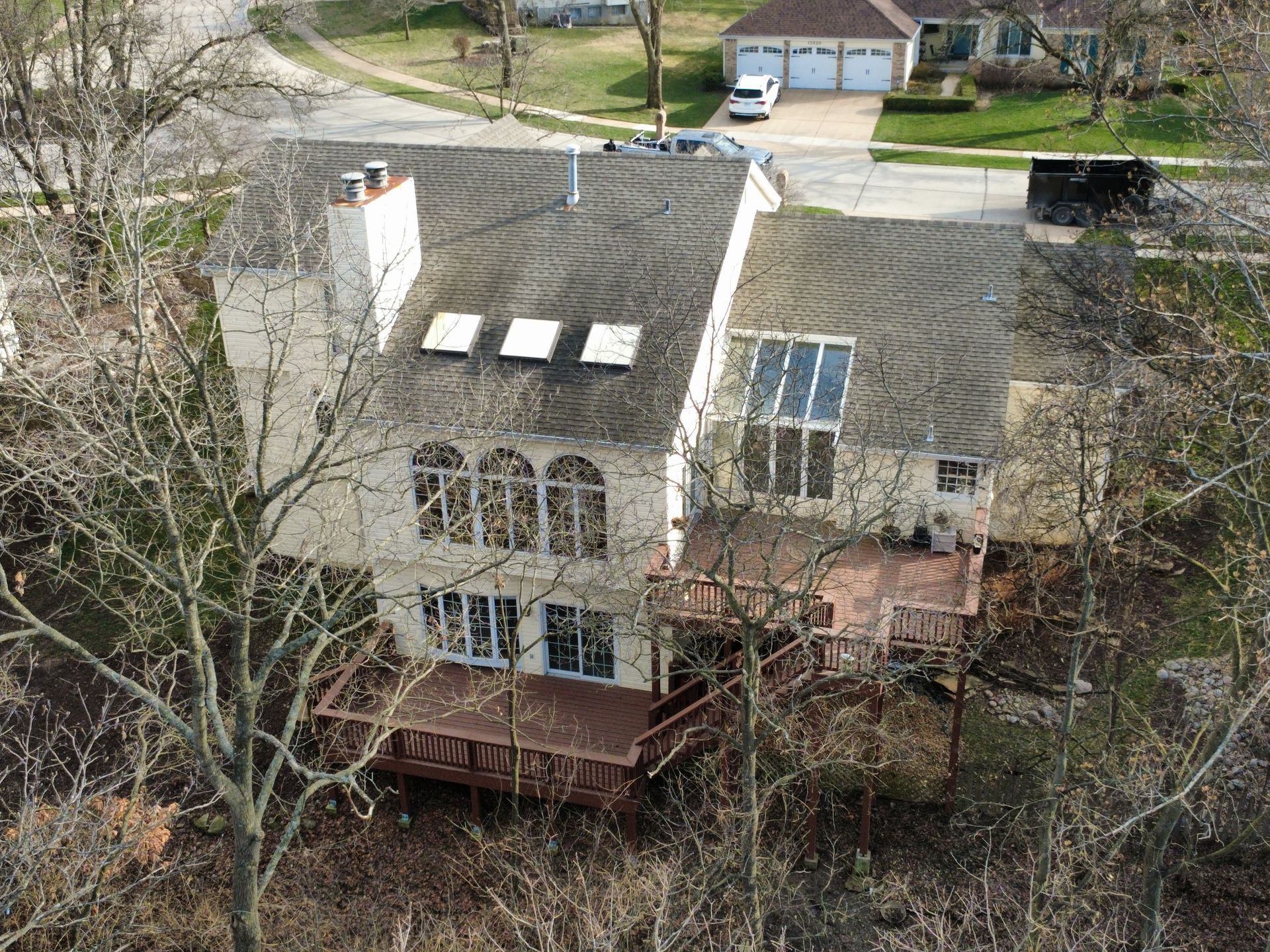 An aerial view of a multi-story home with a light-colored exterior, brown roof, and a wooden deck, set in a wooded area.