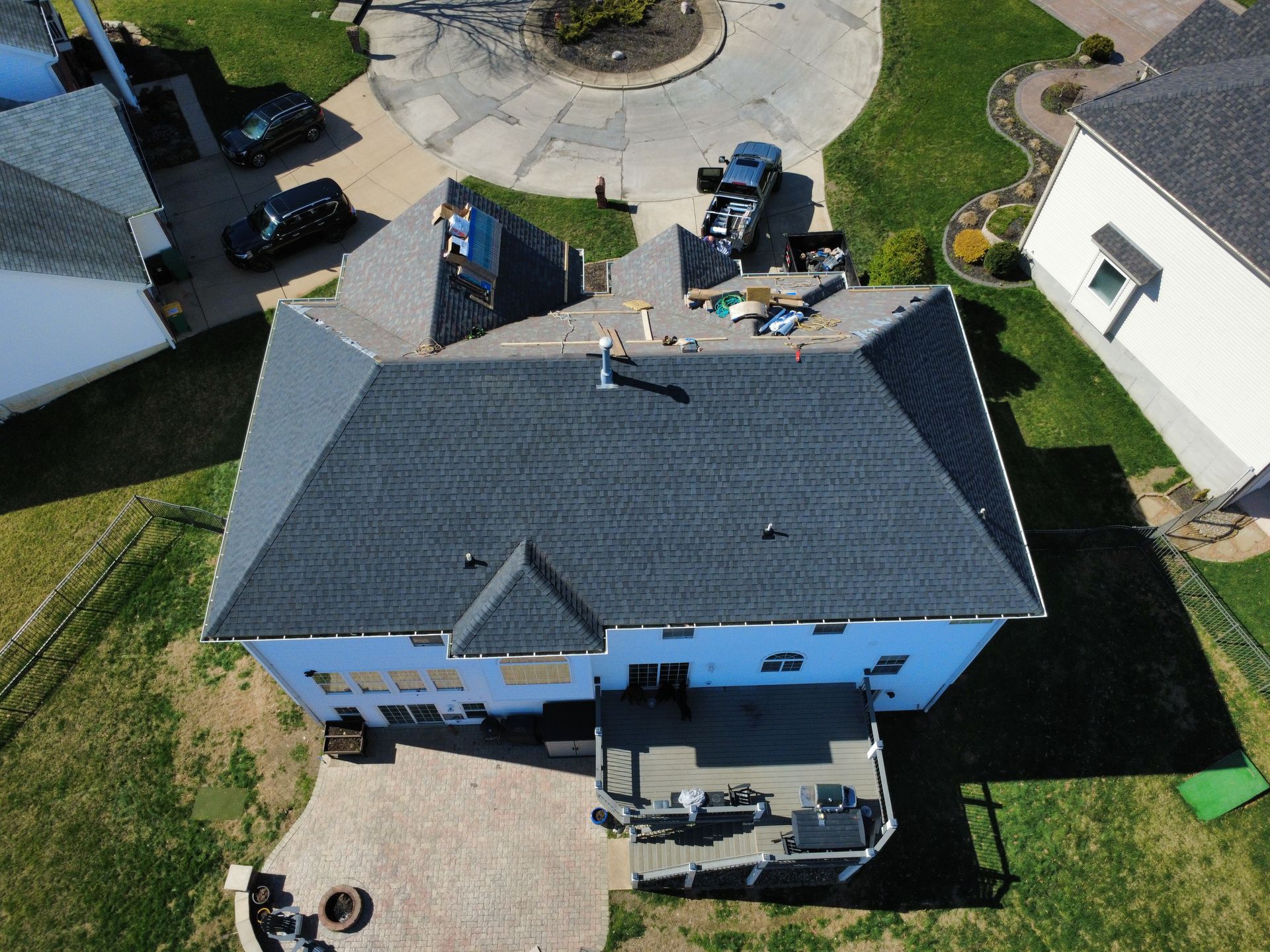 An aerial view of a residential house with a new dark gray shingled roof, showing construction supplies and a backyard.