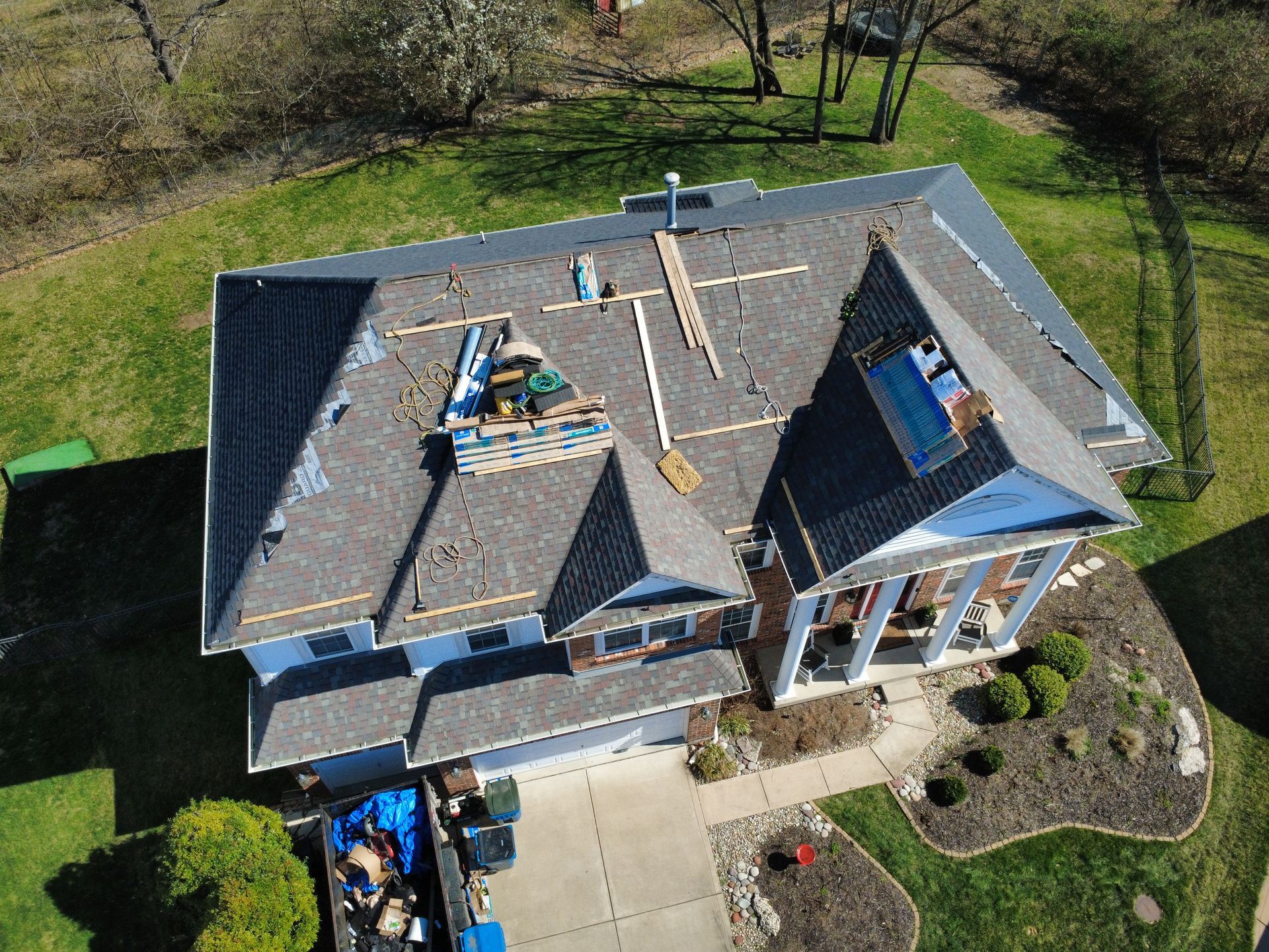 Aerial view of a residential roof undergoing construction, with shingles, tools, and a dumpster in the driveway below.