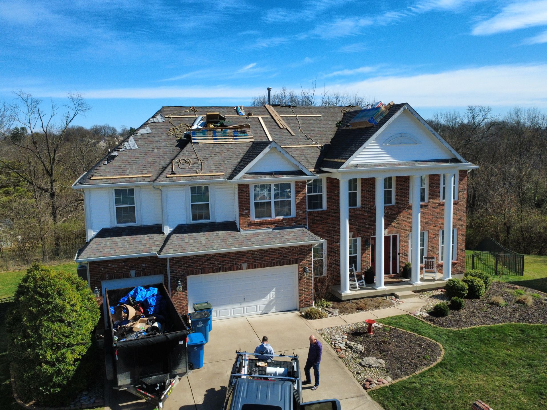 Workers performing roof repairs on a large two-story brick home with a white porch, featuring a dumpster in the driveway.
