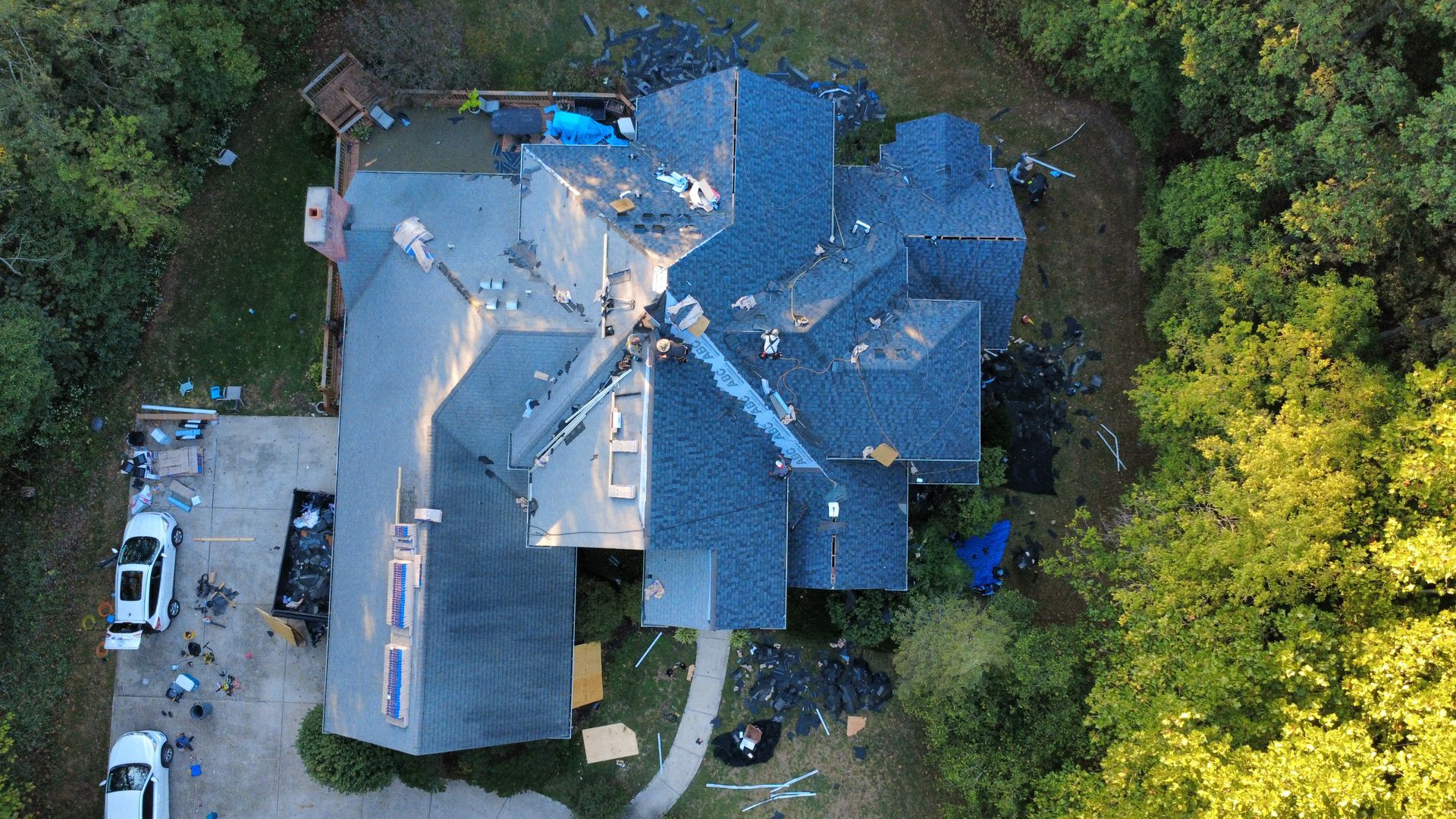 An aerial view of a house undergoing roof repairs, featuring blue shingles, a construction dumpster, and two parked cars.