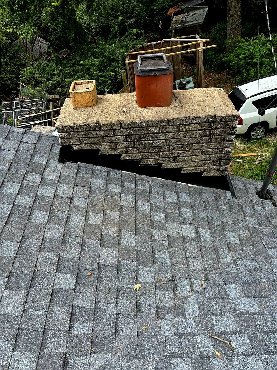 A stone chimney with two flues stands on a shingled roof, showing a significant gap where flashing should be installed.