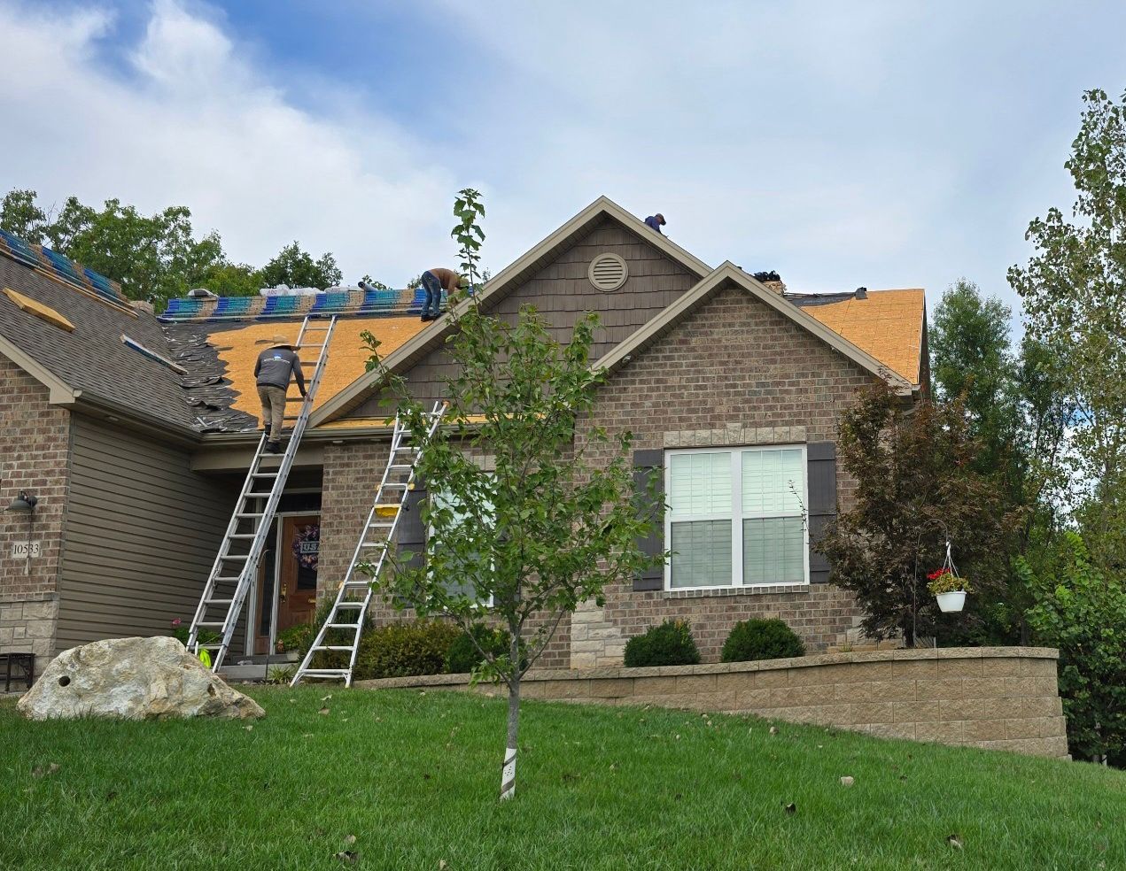 Workers repairing the roof of a brick house with two ladders set up against the front.
