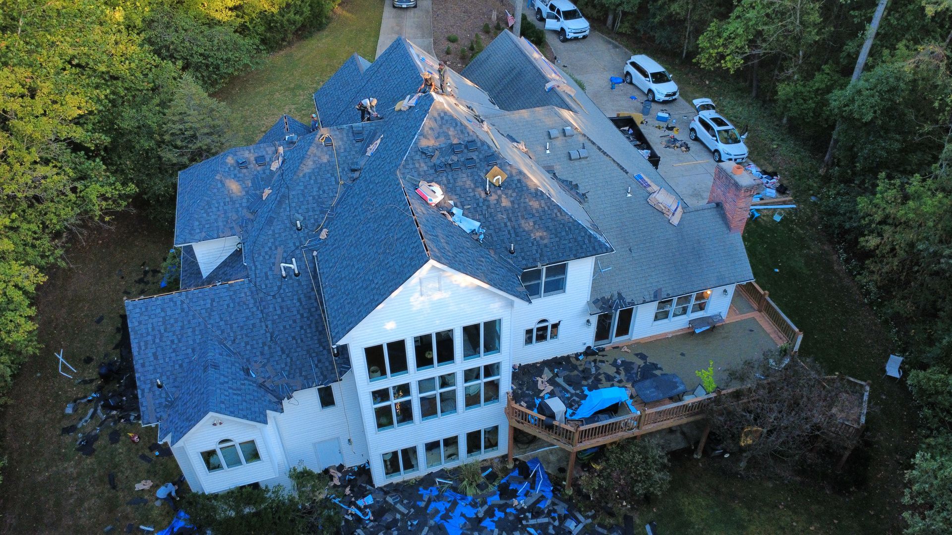 Aerial view of a white multi-story house undergoing roof repairs, with workers on the roof and materials in the yard.