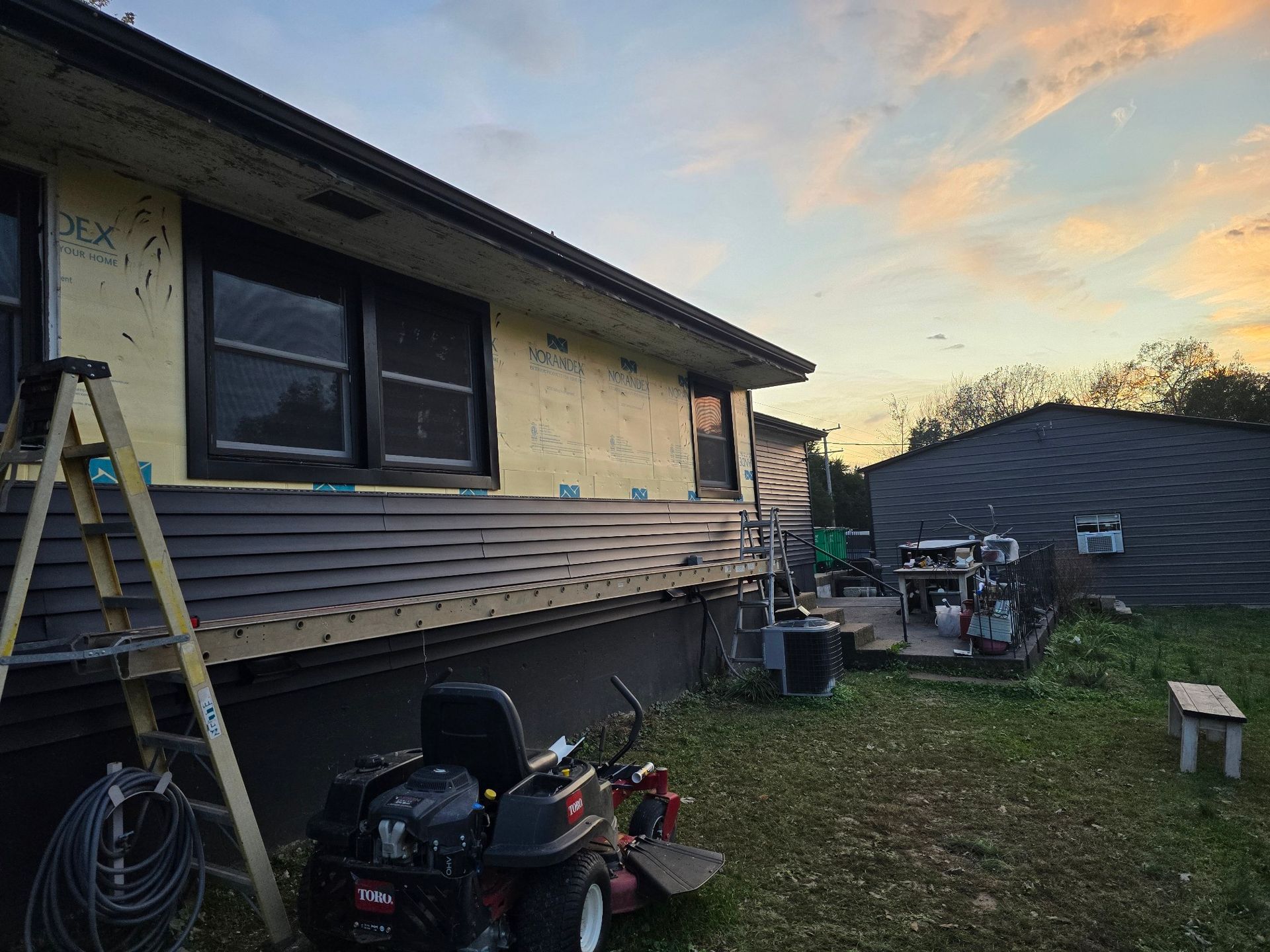A house under renovation with dark horizontal siding, a ladder, a lawn mower, and another building at sunset.
