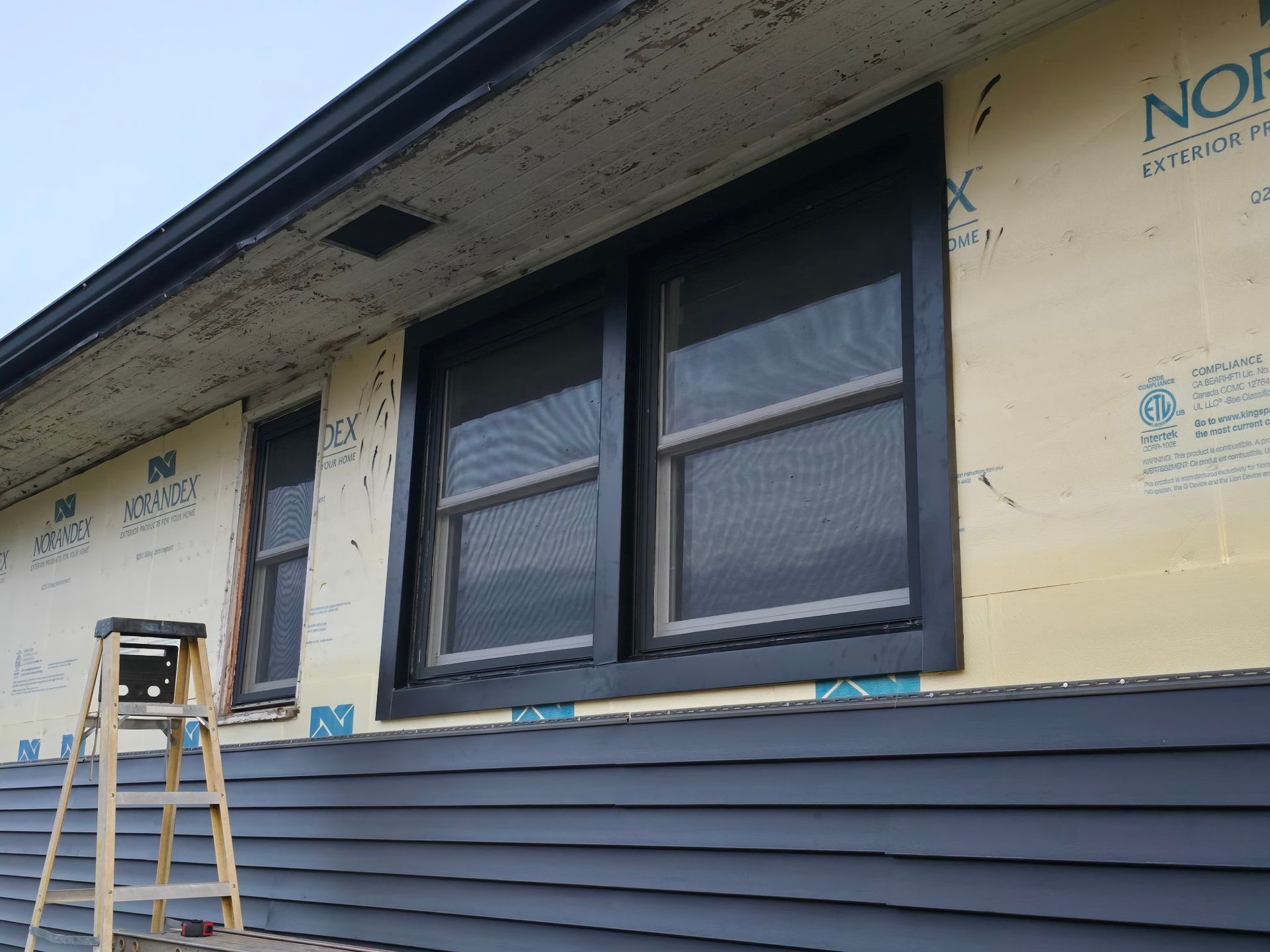 An unfinished exterior wall with dark siding, black-trimmed windows, yellow insulation board, and a stepladder.