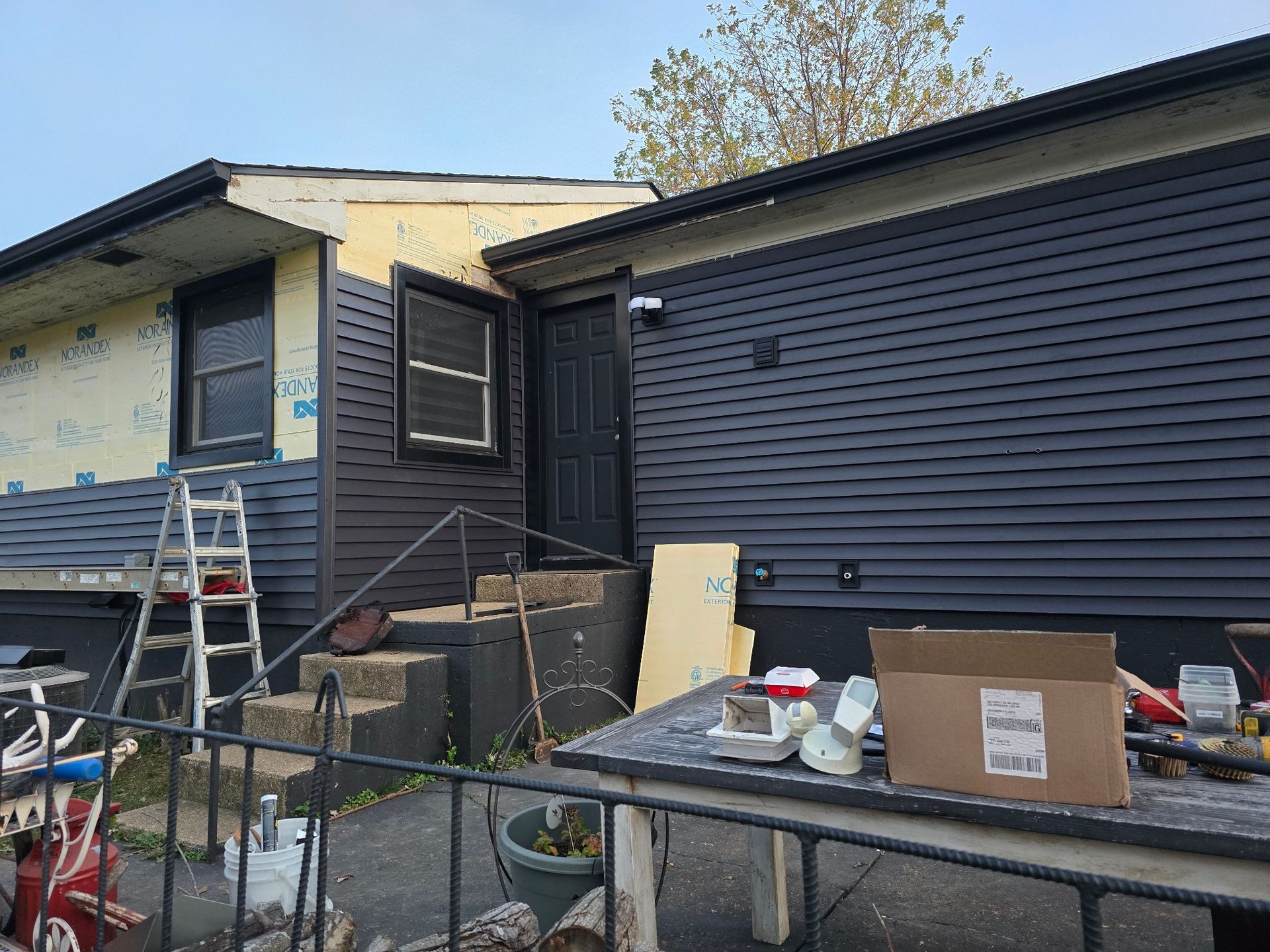 A house exterior under construction with dark siding partially installed, a ladder, and a table with supplies.