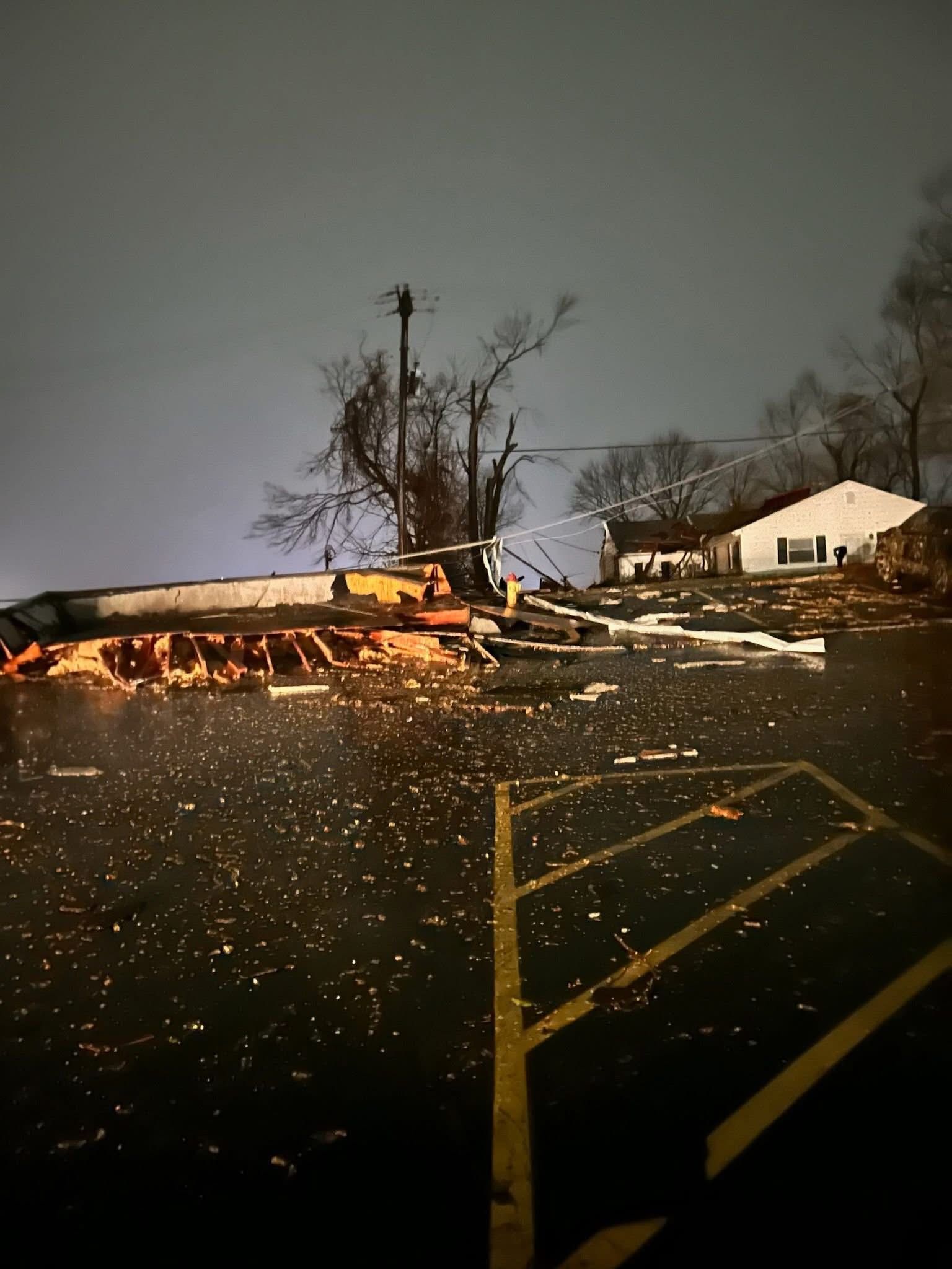 A damaged building with debris scattered across a parking lot at night, beneath a dark, cloudy sky.