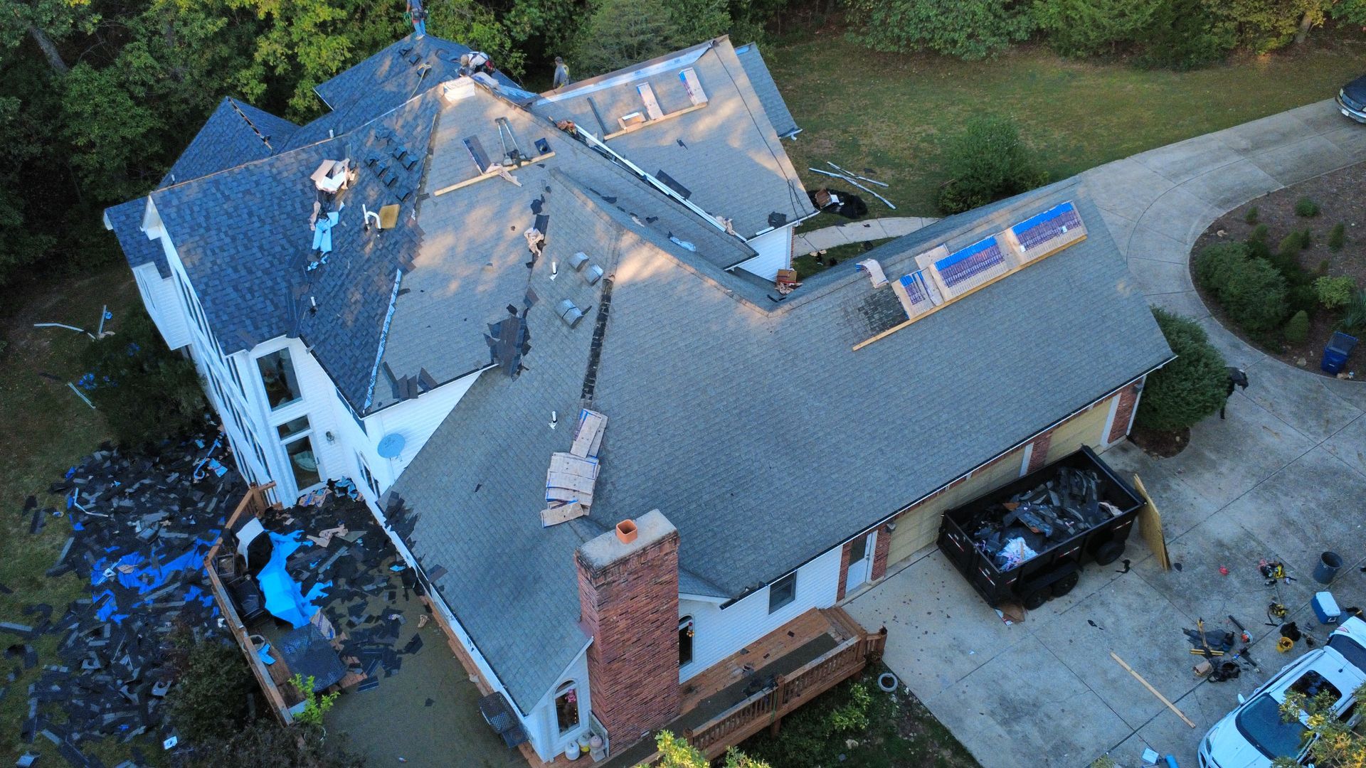 Aerial view of a residential house undergoing roof replacement, with piles of old shingles and a dumpster on the driveway.