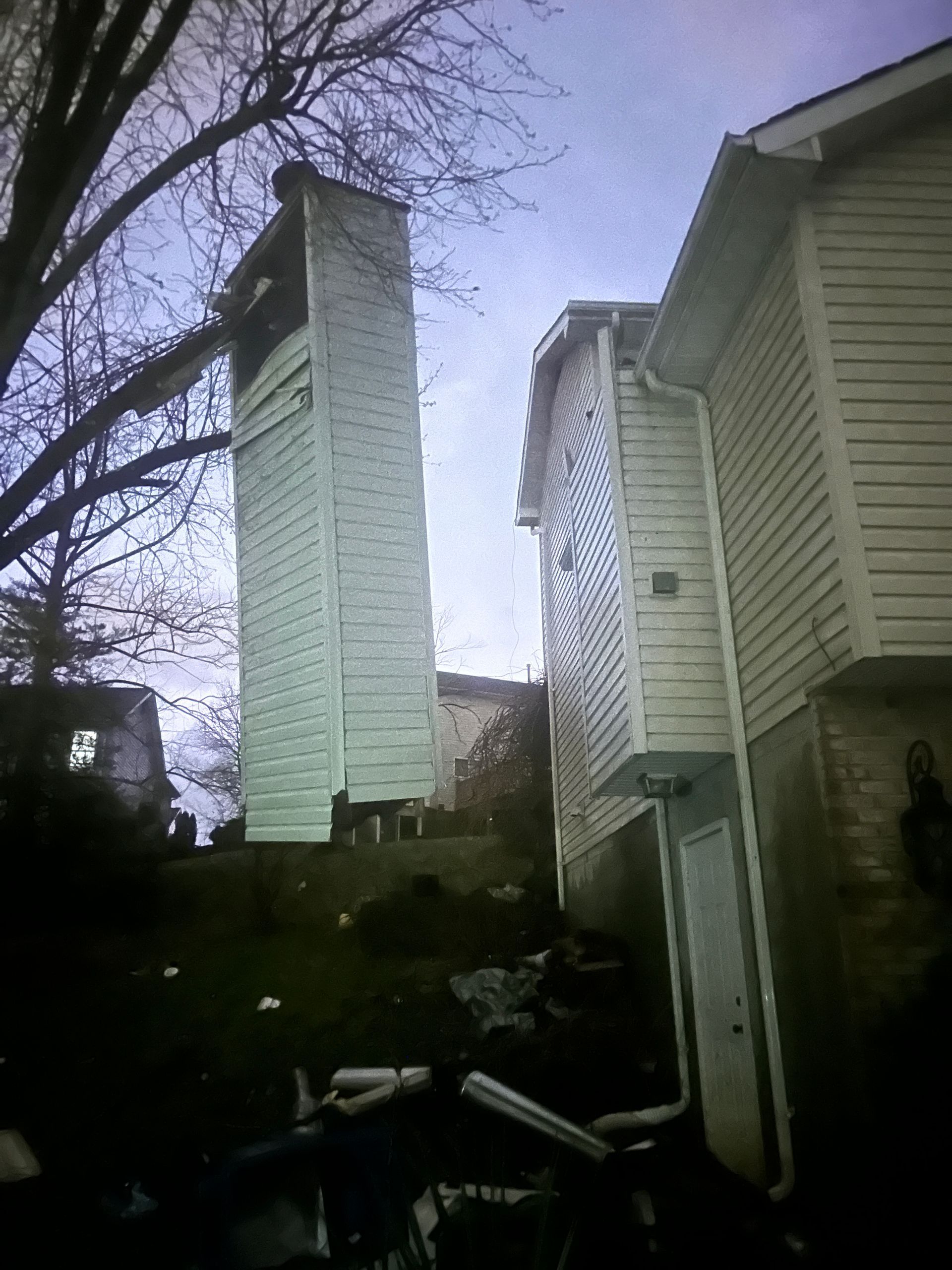 A detached, narrow chimney structure stands upright in a yard next to a beige residential house with vinyl siding.