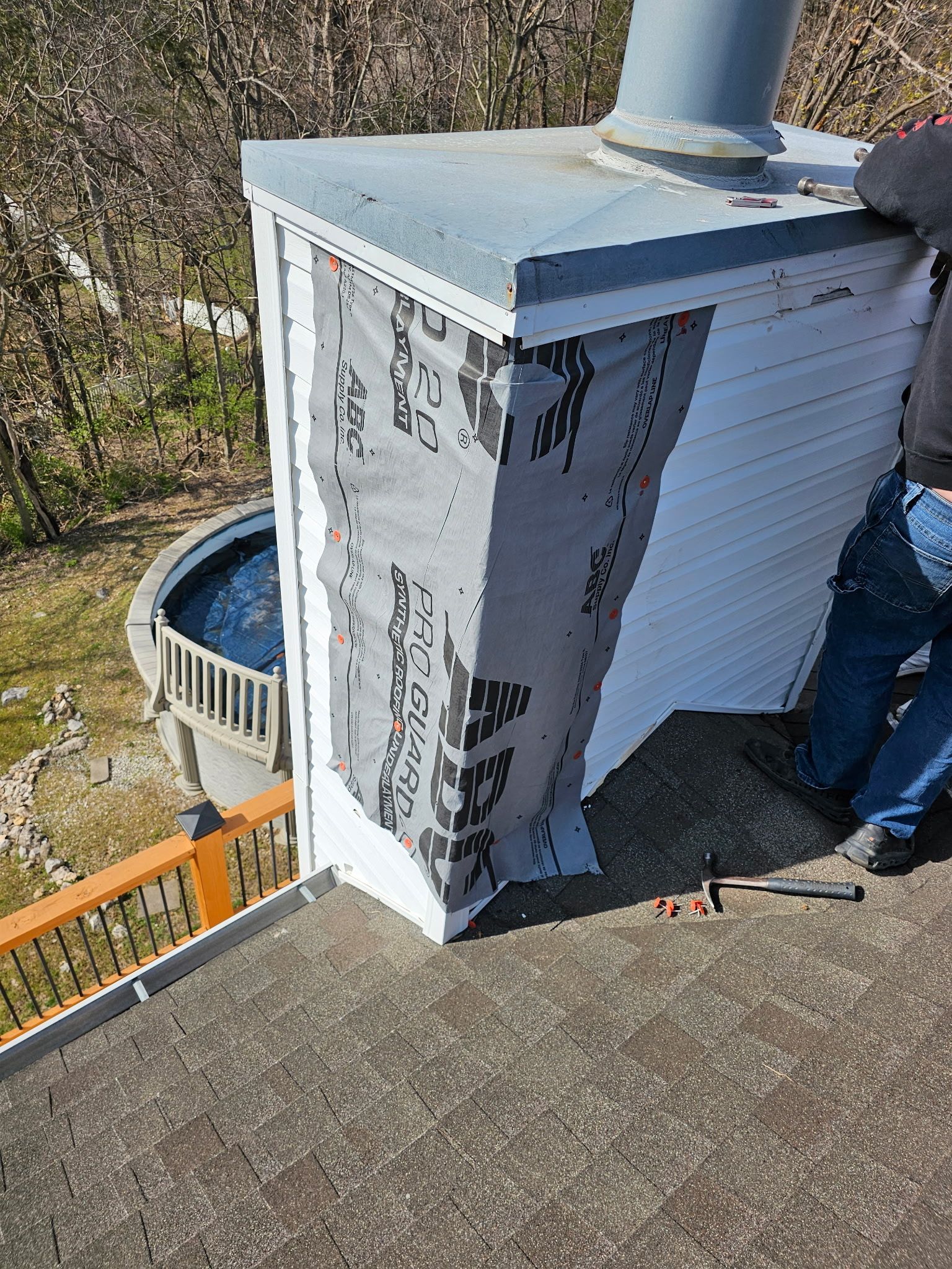 A worker applies grey flashing material to the corner of a white chimney on a shingled roof.