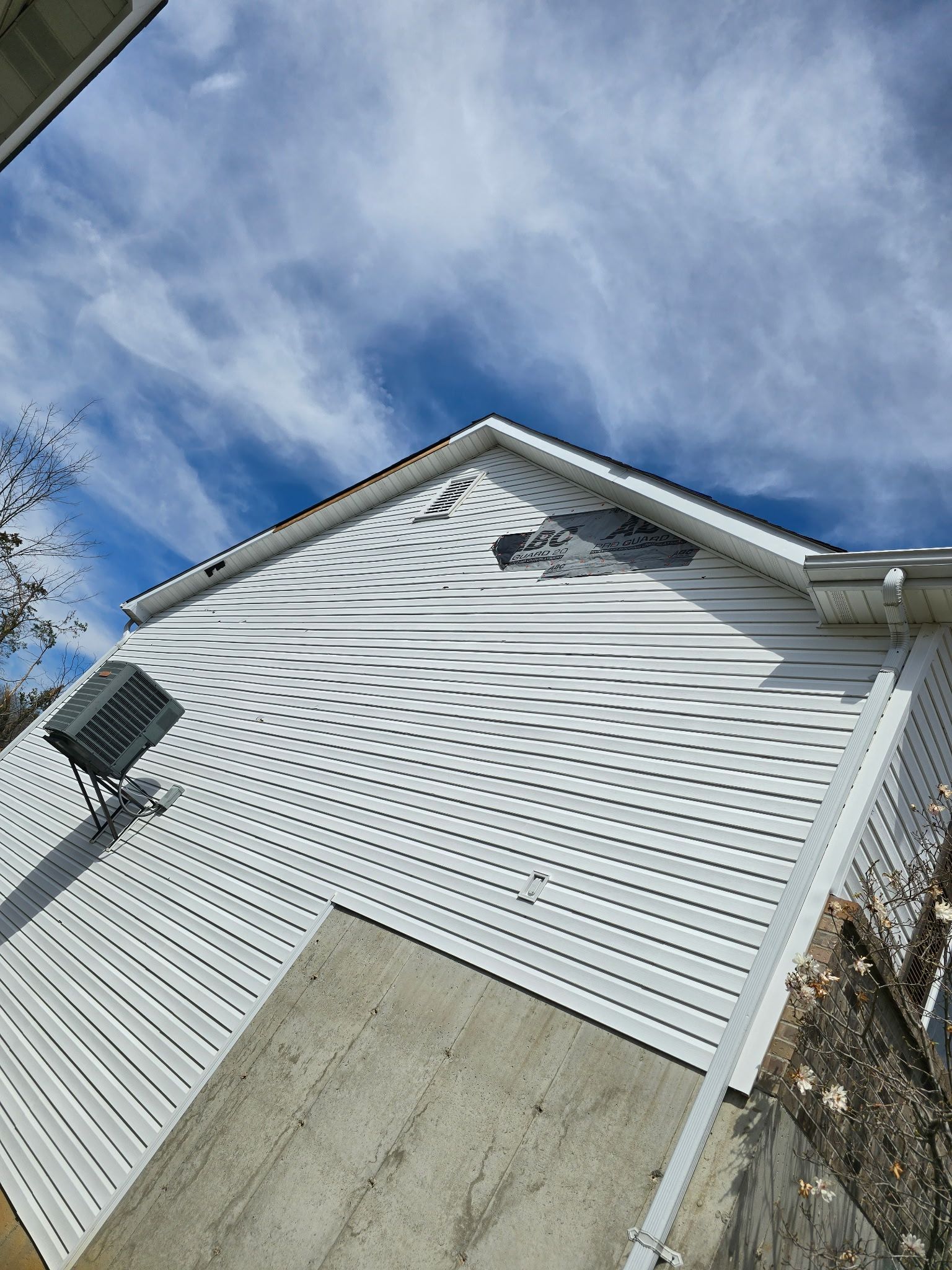 A triangular wall with white horizontal siding and a moiré shadow pattern cast by a nearby fence under a blue cloudy sky.