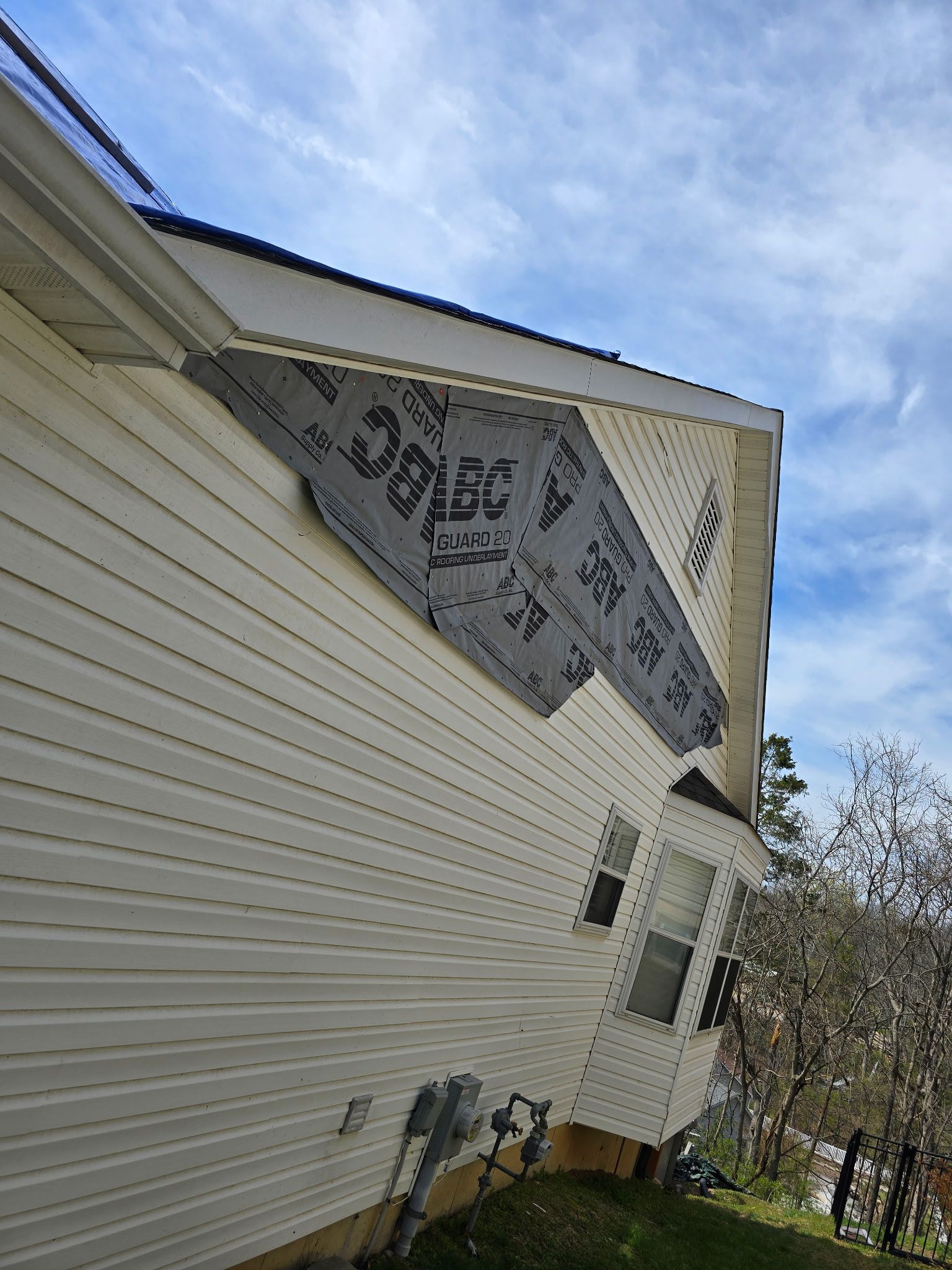 A house exterior shows missing vinyl siding on a gable, revealing grey house wrap and wood sheathing underneath.