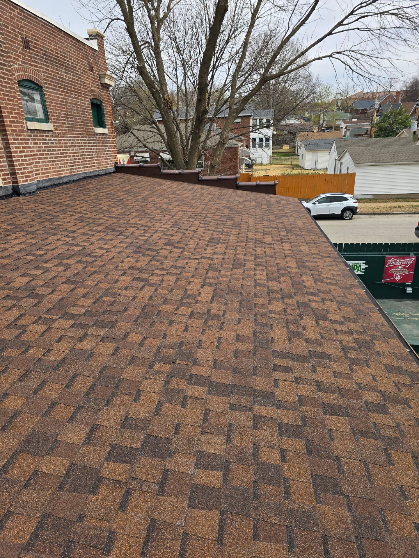 A view from a rooftop featuring brown asphalt shingles, with a brick building wall, trees, and a street scene below.